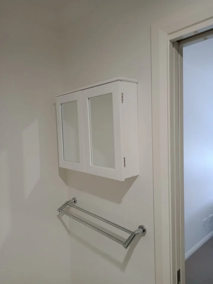 A white wall-mounted cabinet with glass doors above a chrome towel bar, near a doorway with white trim.