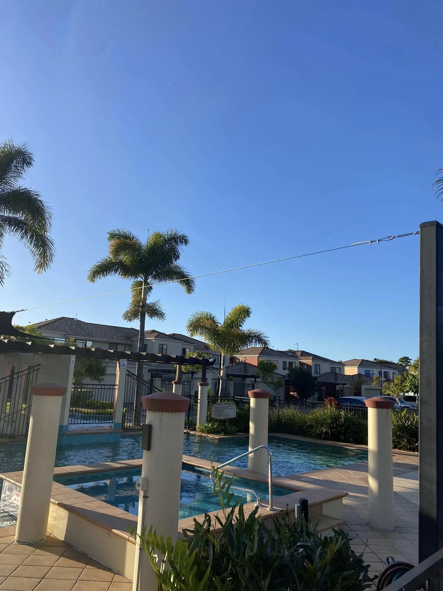 Empty outdoor hot tub with a fenced pool area, surrounded by palm trees and residential buildings, under a clear blue sky.
