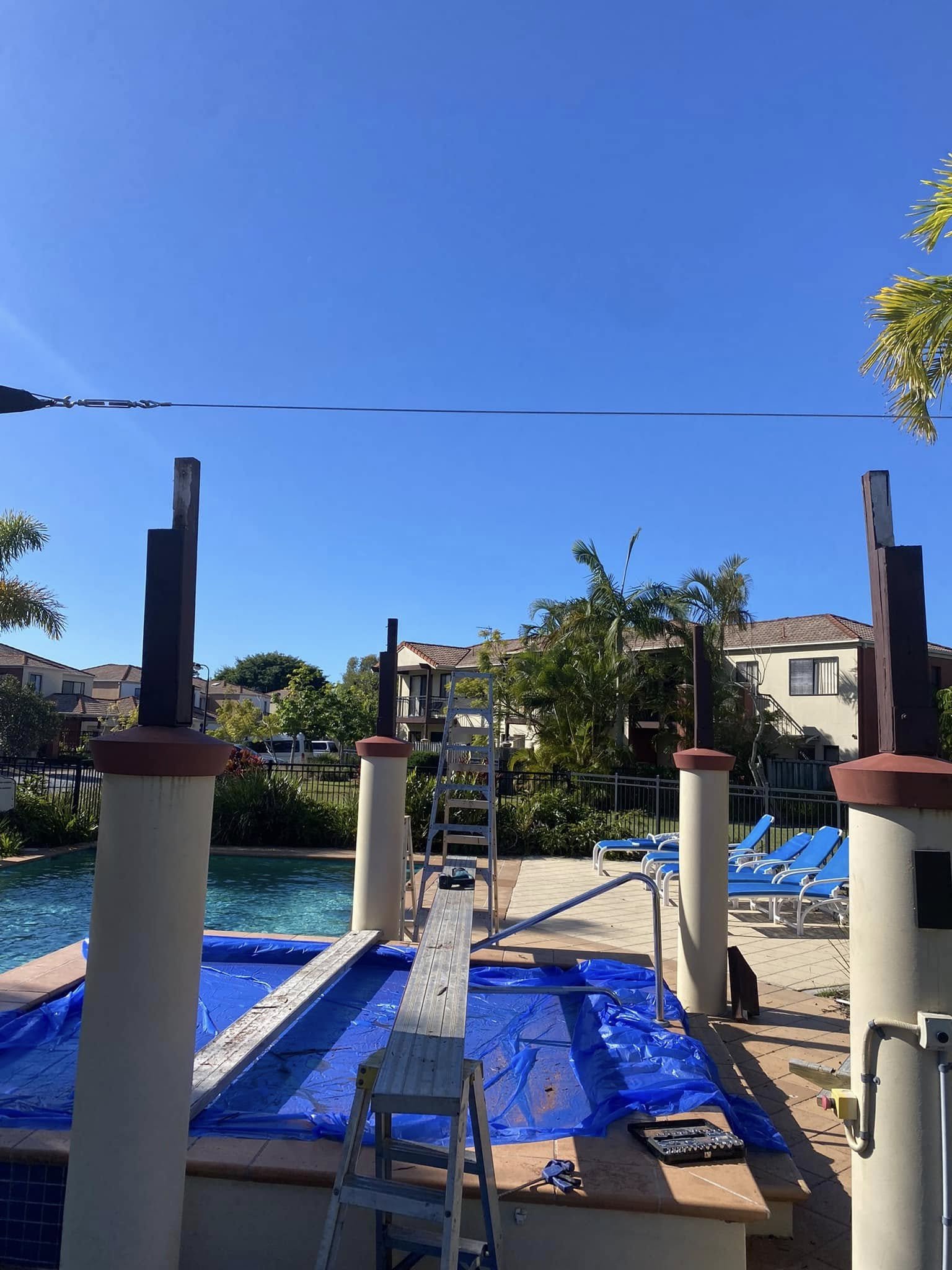 Swimming pool area under repair with a ladder, blue pool cover, and lounge chairs, surrounded by residential buildings and palm trees.