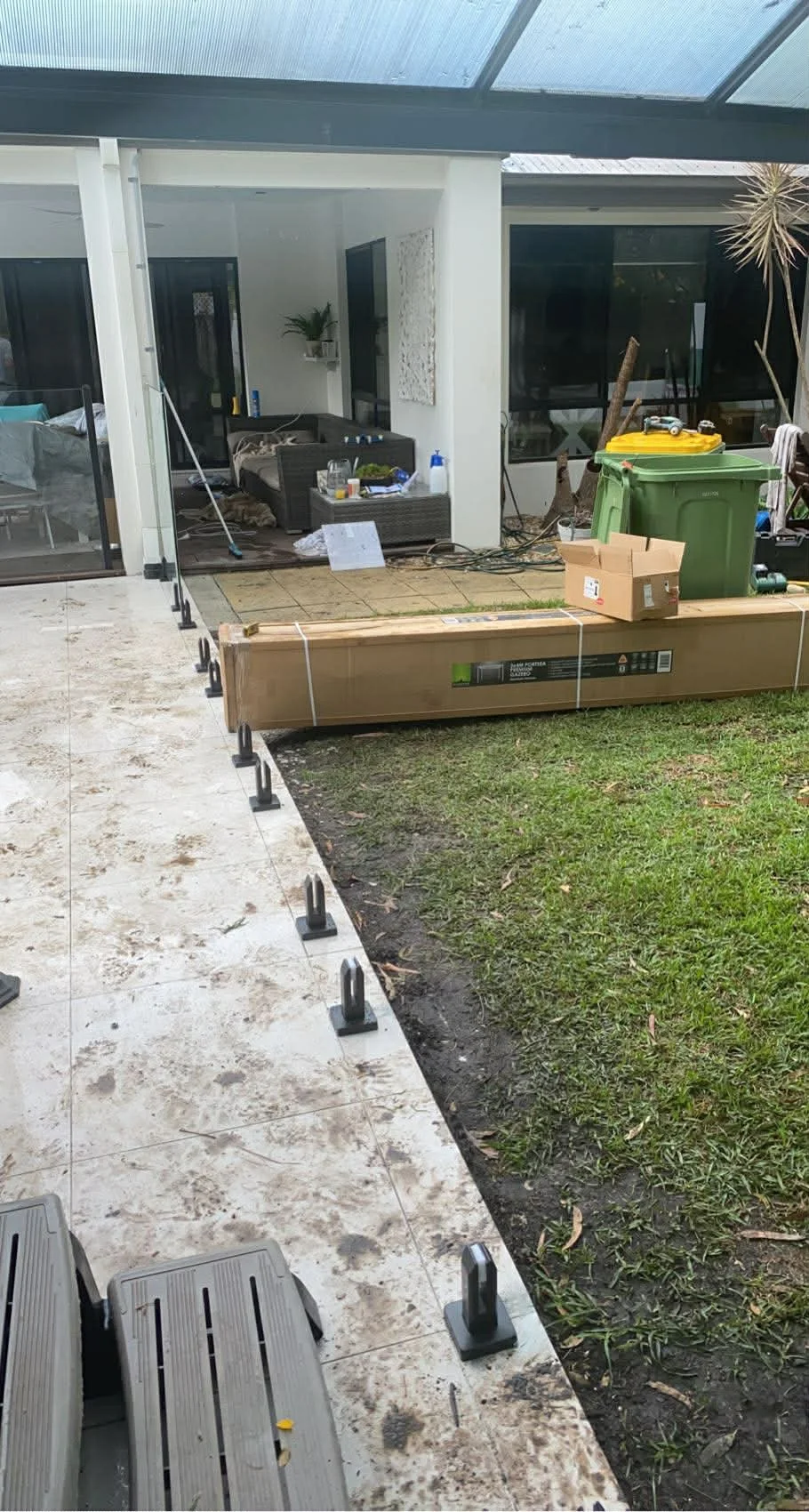 Backyard under renovation with tiles being installed, construction tools and materials, a green trash bin, a cardboard box, and a partially completed patio area.