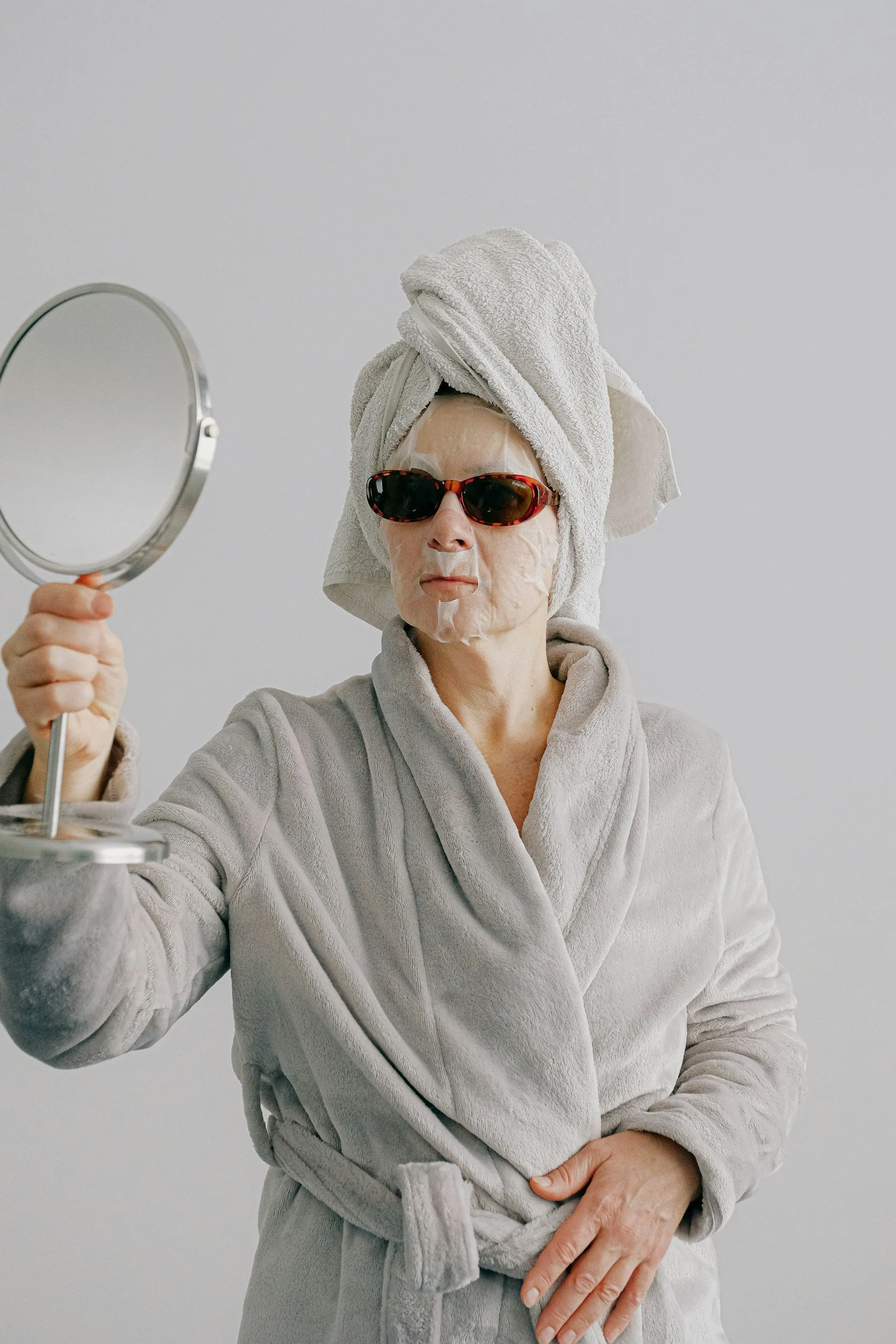 Innere Klarheit: Frau in Bademantel mit Handtuch im Haar, Sonnenbrille, schaut in den Spiegel, bei Schönheitsroutine und dem Blick nach innen.