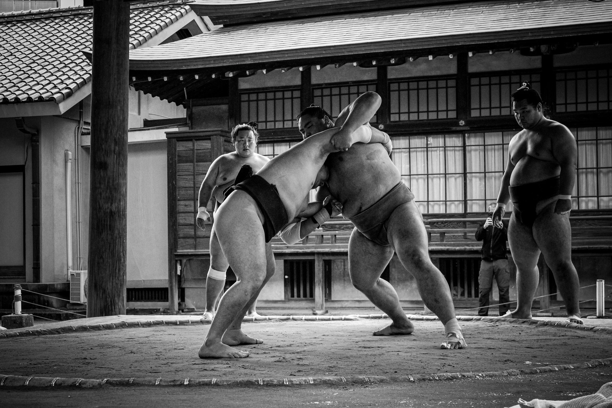 Starkes Miteinander: Zwei Sumo-Ringer kämpfen in einem Ring, während drei weitere Beobachter zuschauen.