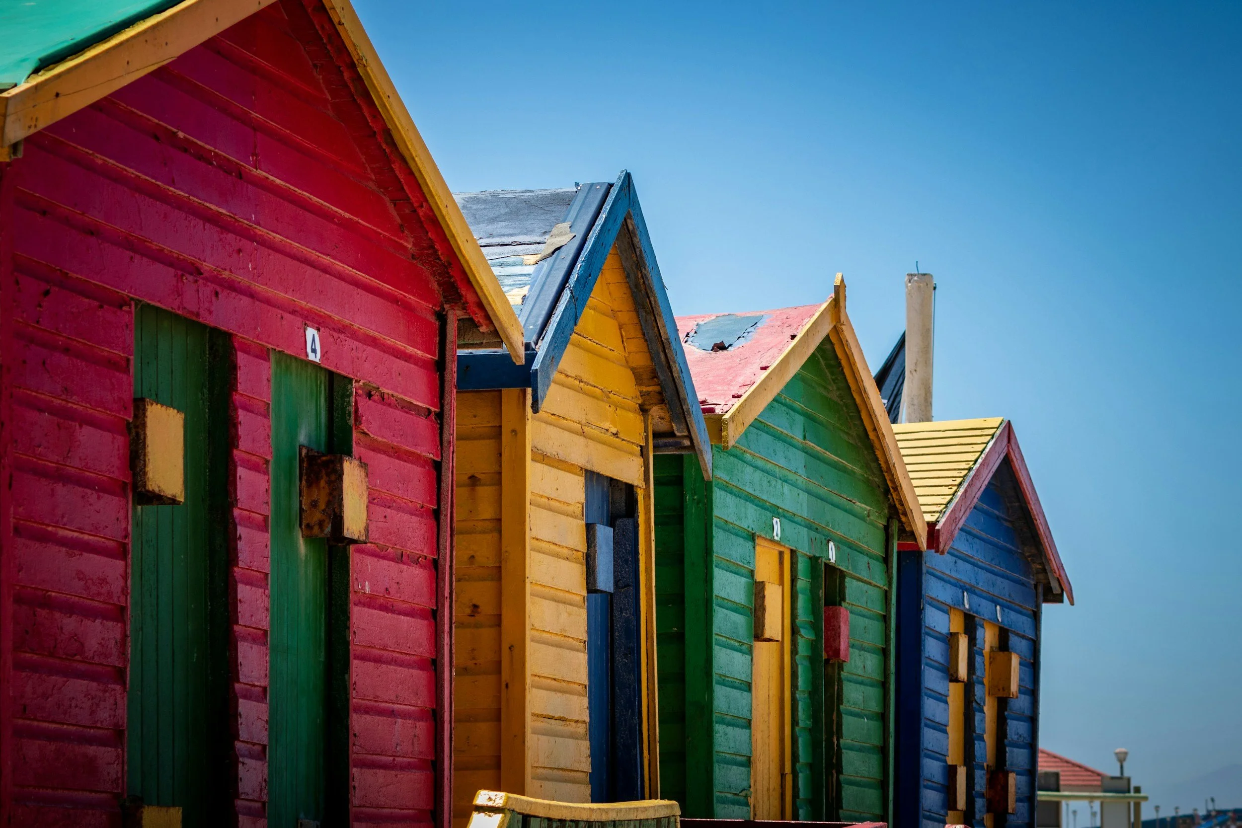 Bunte bemalte Strandhütten mit roten, gelben, grünen und blauen Wänden und Dächern, aufgenommen bei sonnigem Wetter gegen den blauen Himmel.