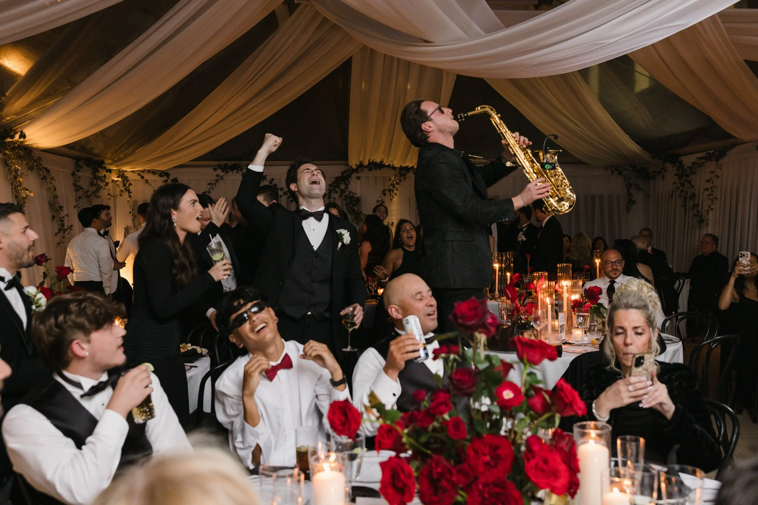 People at a festive wedding dinner in dancing, watching Jason Nelson play live, high-energy saxophone in an elegant decorated venue with draped fabric ceiling, floral arrangements, and candles.