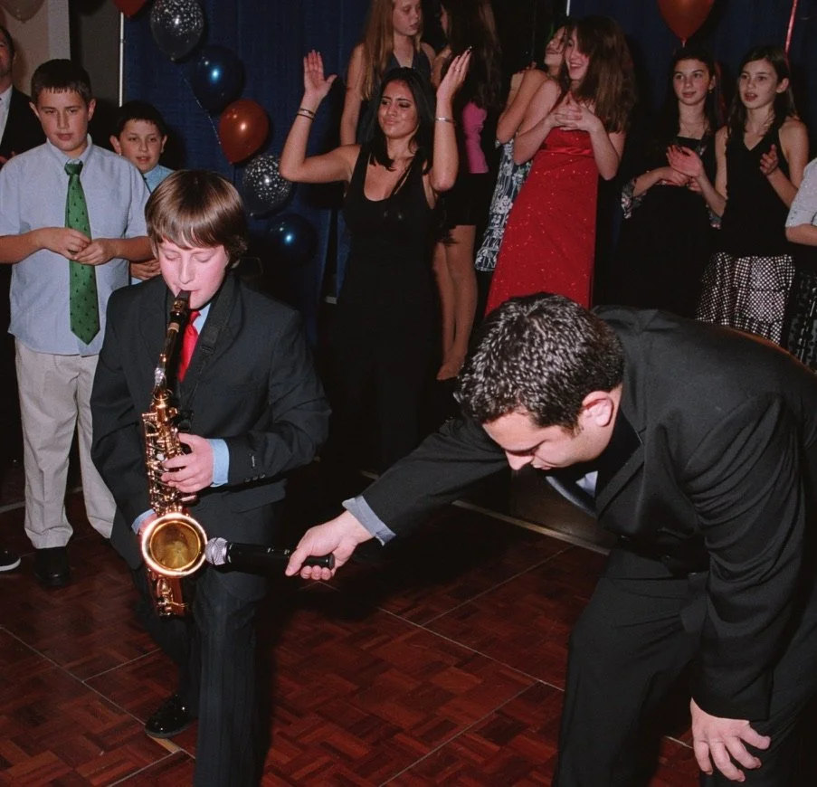 Young Jason Nelson in a suit playing the saxophone at a bar mitzvah with a group of people dancing and socializing in the background.
