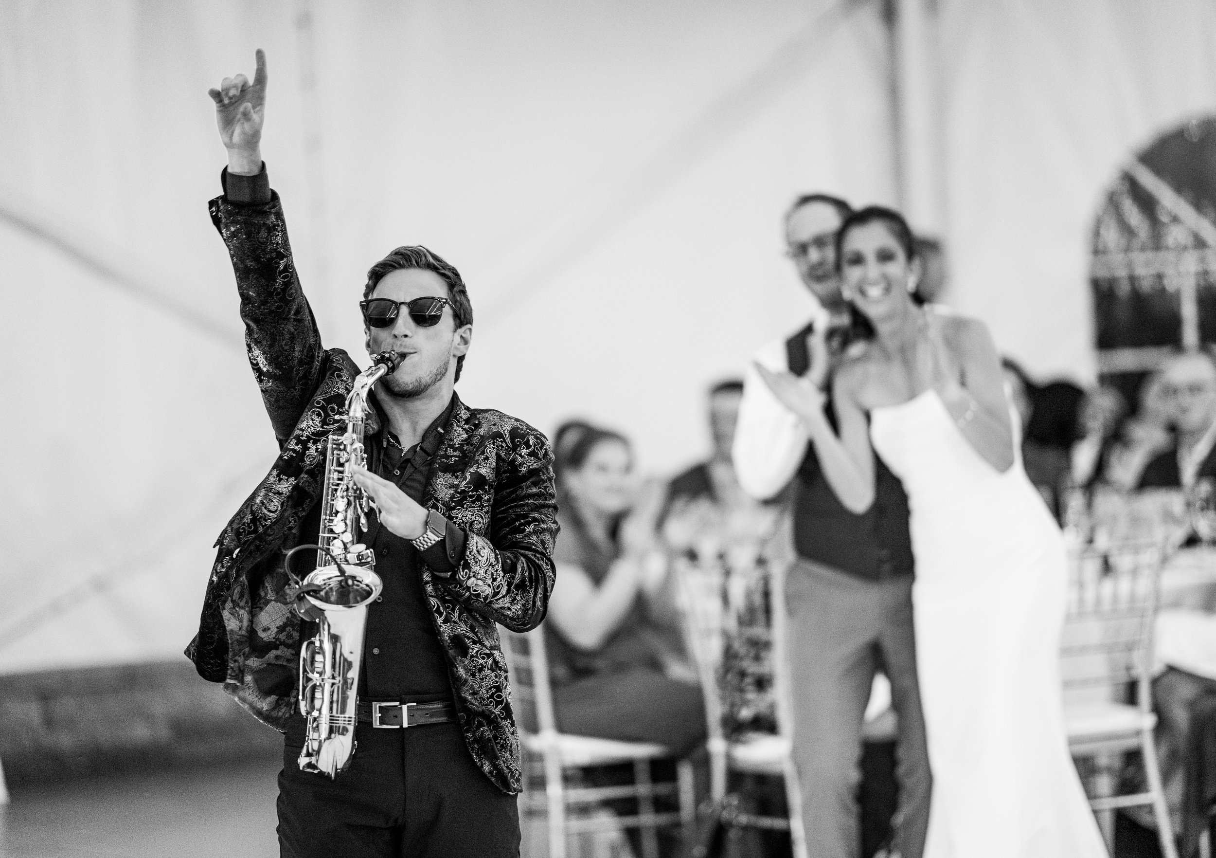 Jason Nelson wearing sunglasses plays a saxophone, with a woman in a dress clapping in the background, in an indoor setting.