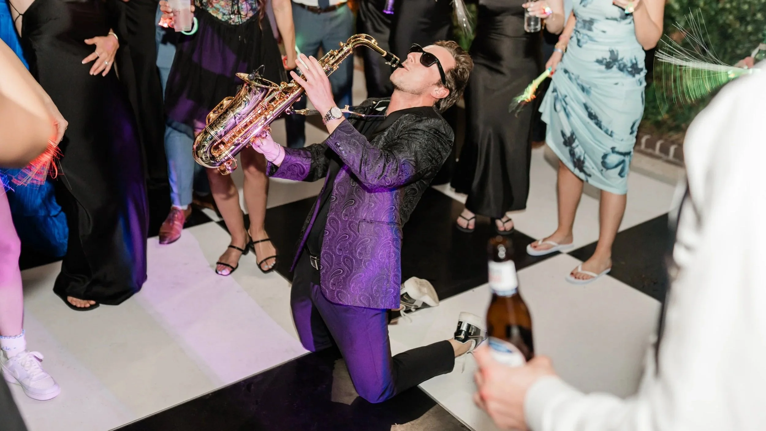 Jason Nelson in a purple suit playing a saxophone on his knees at a club in Los Angeles with people dancing around him.
