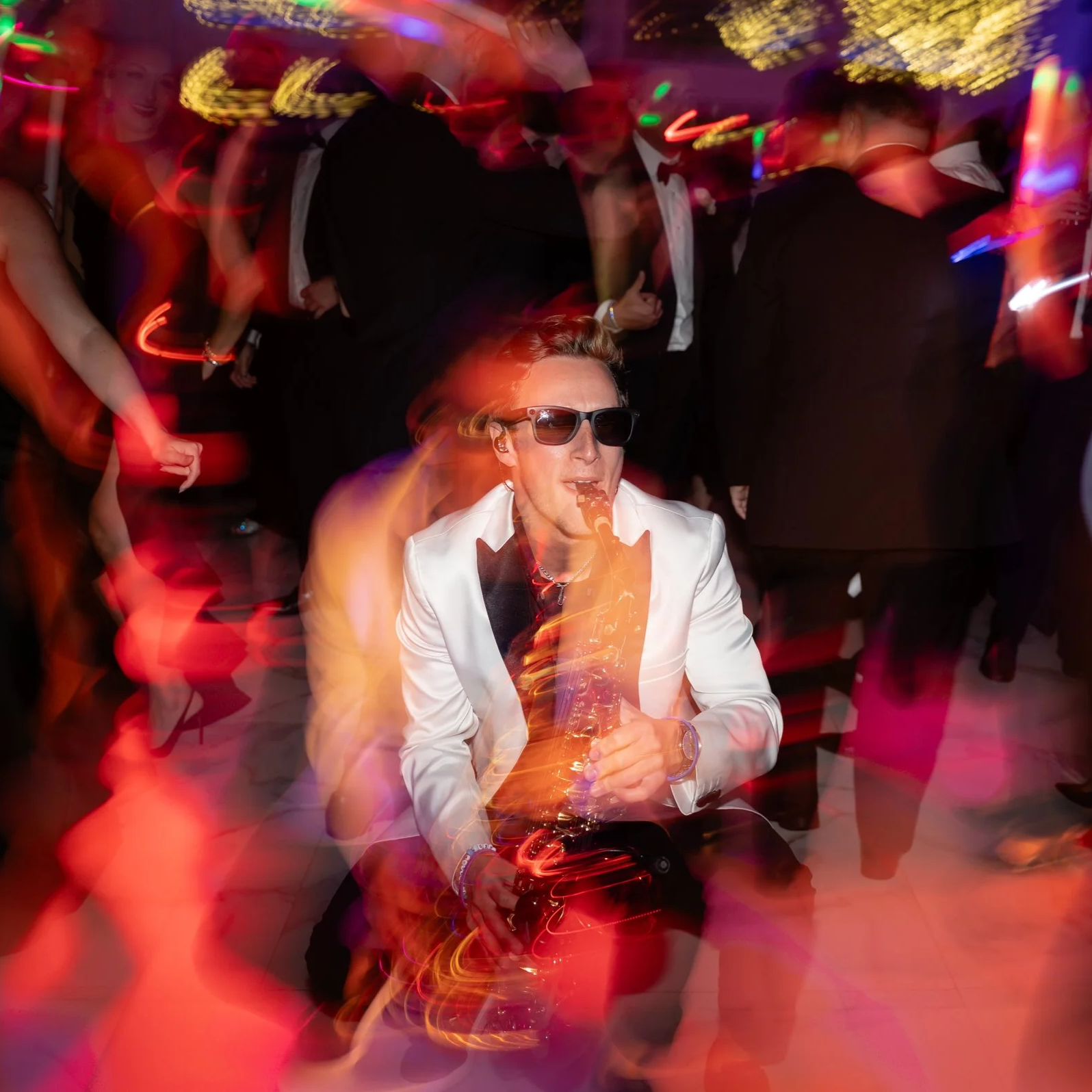 Jason Nelson wearing sunglasses and a white suit jacket playing a saxophone at a private party with colorful, blurred lights and dancing people in the background.