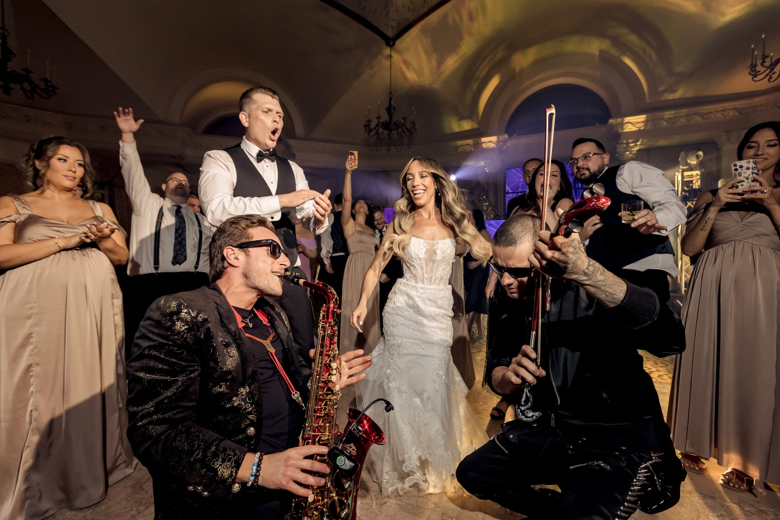 A group of people dancing and enjoying music at a wedding reception in an elegant venue with chandeliers and ornate ceiling. The bride, in a white wedding dress, is smiling and interacting with Jason Nelson, the saxophonist.