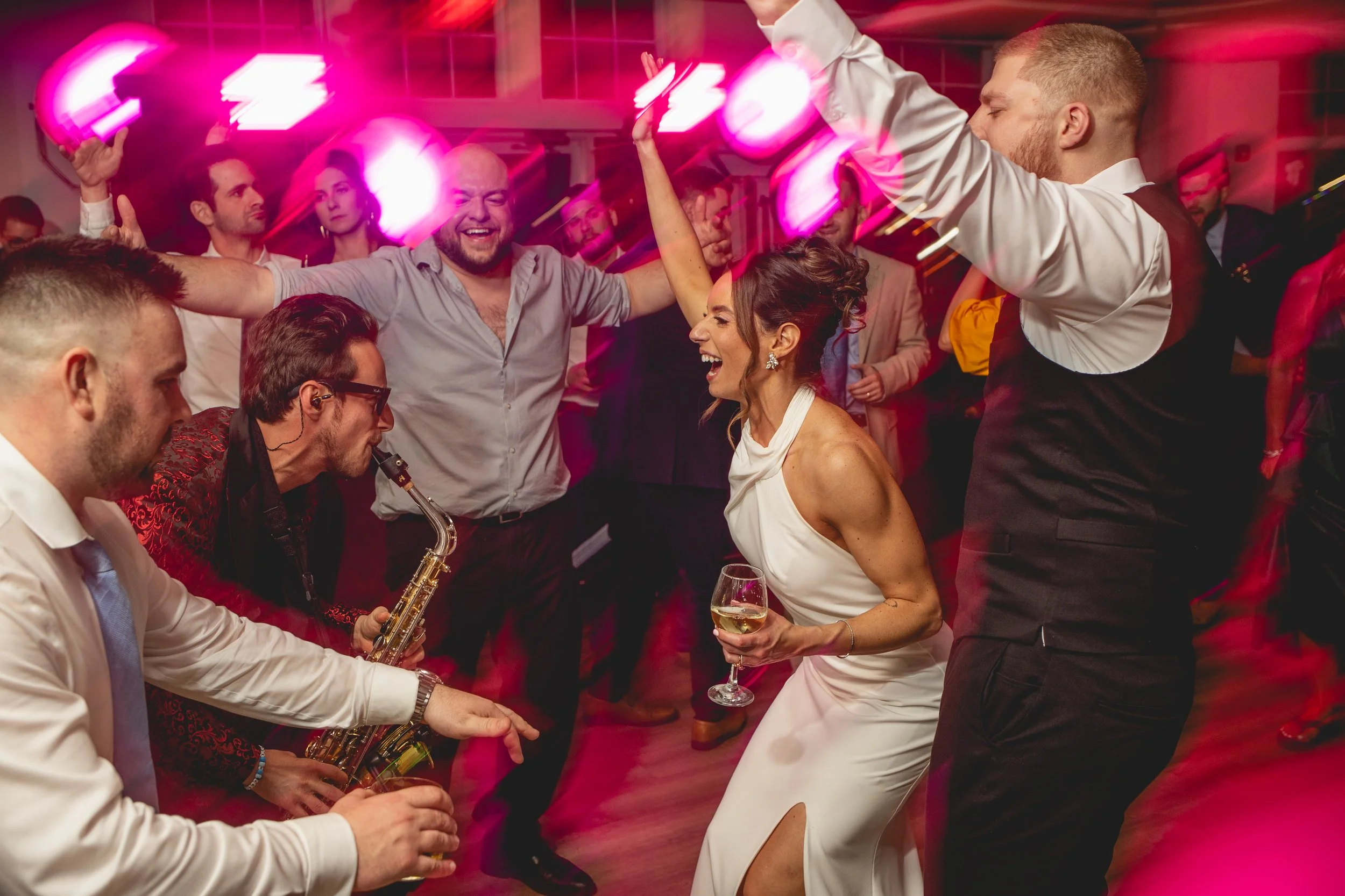 Bride and groom dancing and enjoying music at a wedding with pink and purple lighting, some holding drinks, and a saxophone being played by Jason Nelson.
