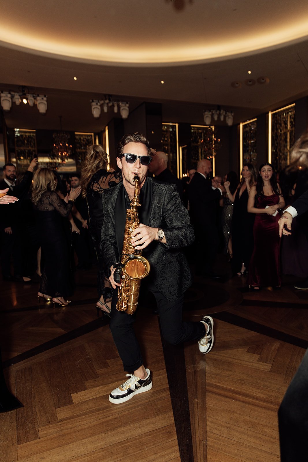 Jason Nelson wearing sunglasses and a patterned blazer playing a saxophone at a NYC wedding reception with guests dancing and socializing in the background.