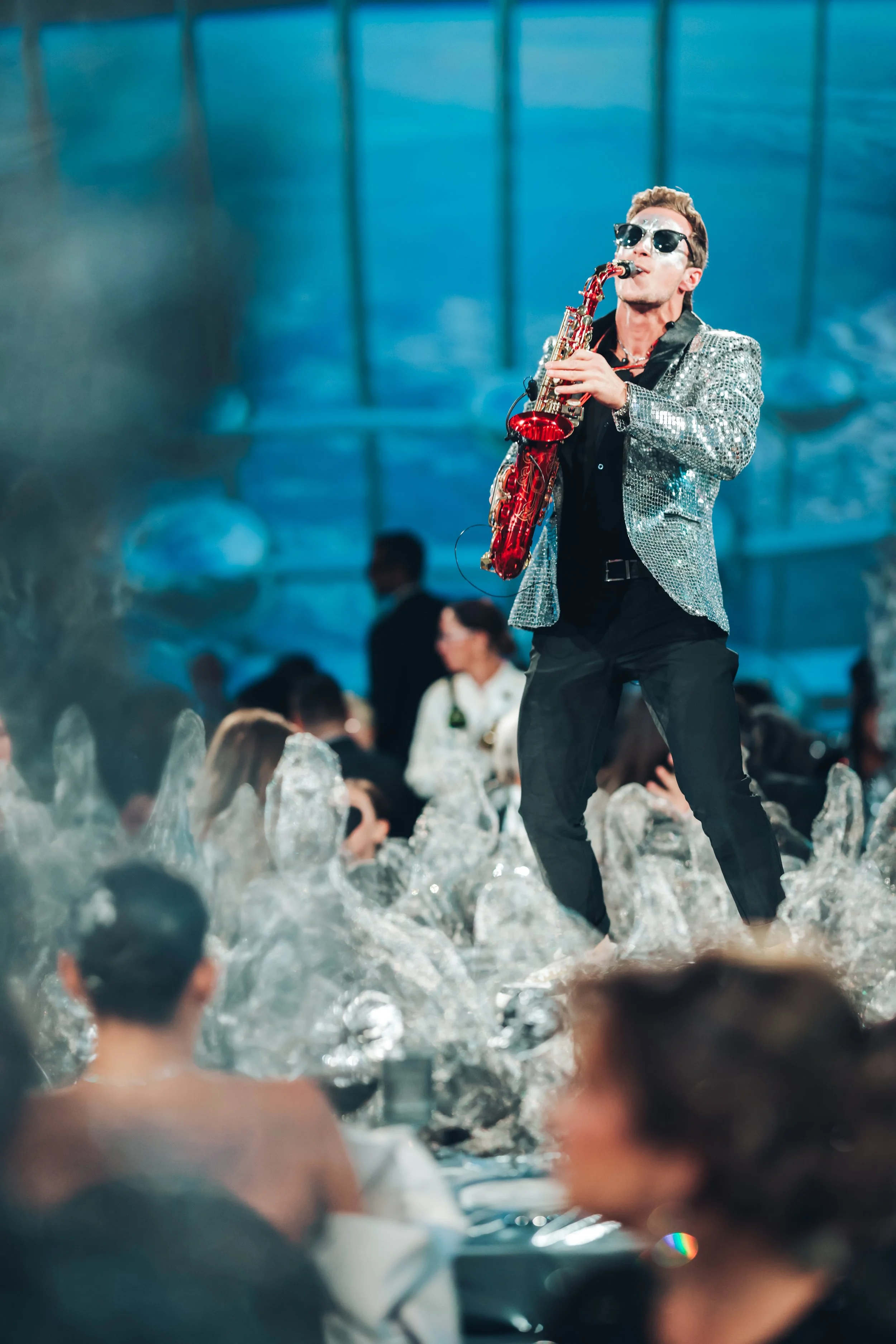 Jason Nelson in a sparkly jacket and sunglasses plays an alto saxophone on stage in front of a corporate party, with colorful lighting and a blurry crowd in the background.