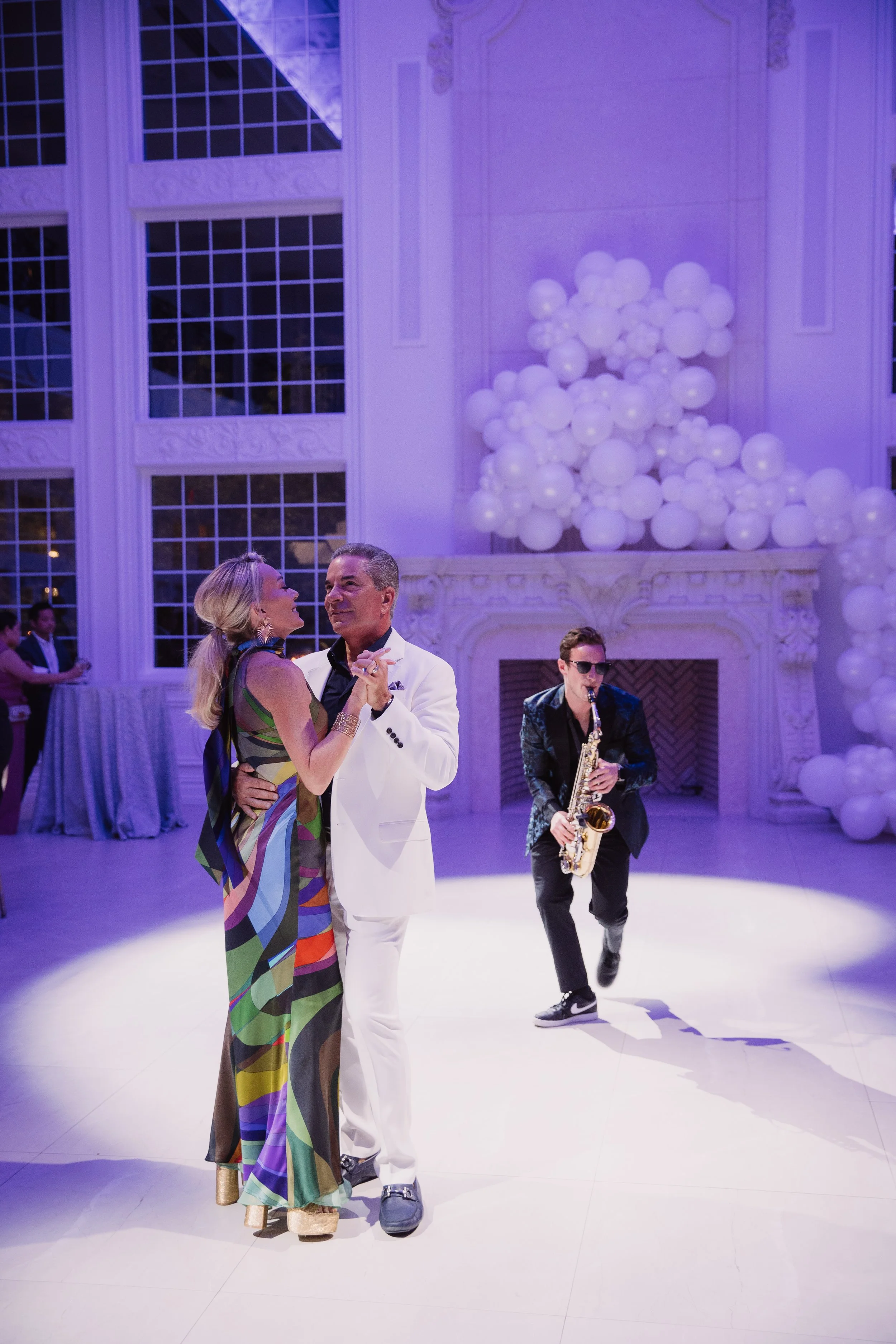 Jason Nelson in a white tuxedo plays live saxophone with a couple dancing in a brightly lit celebration hall decorated with an elaborate balloon display.