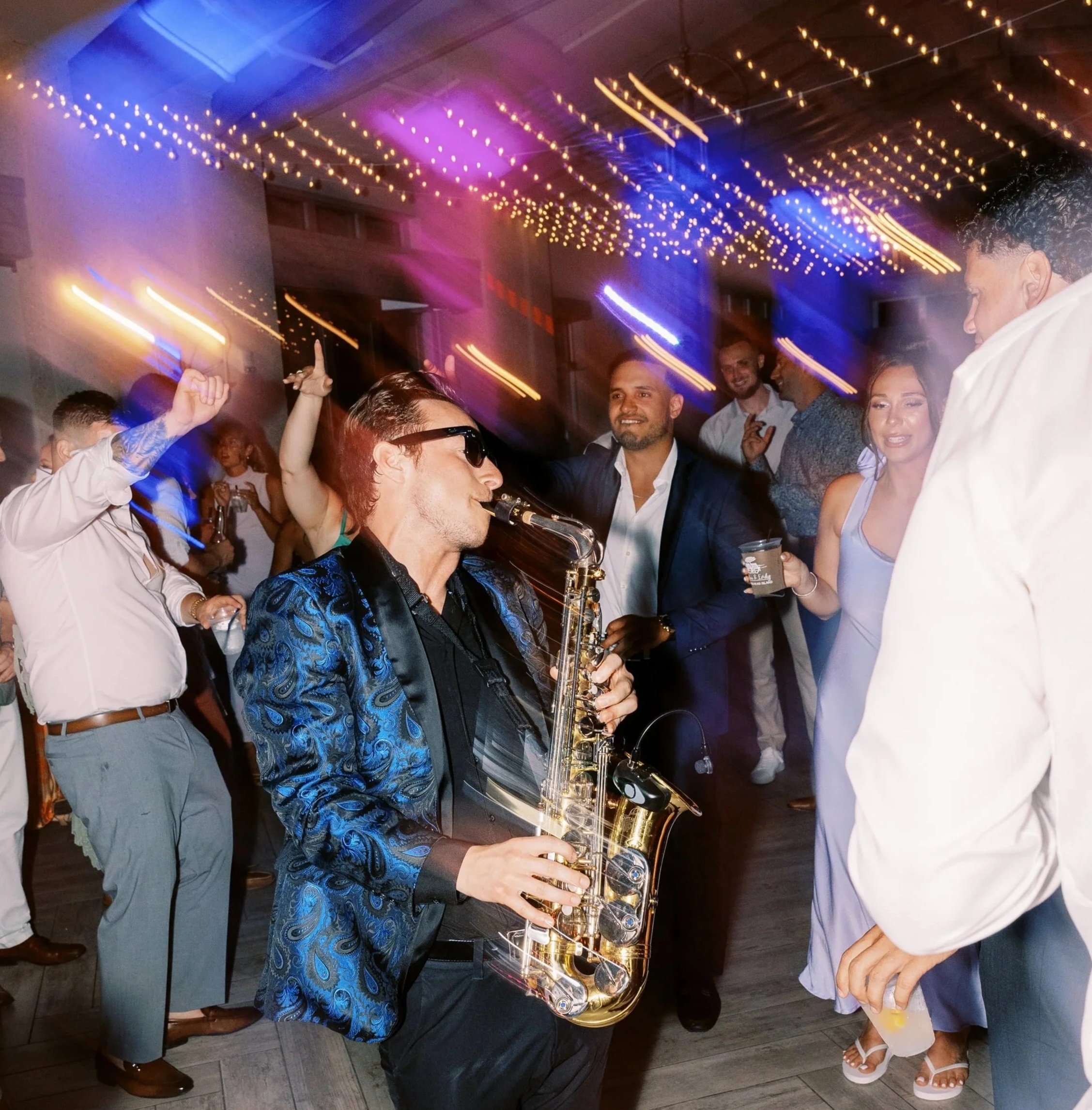 Jason Nelson playing the saxophone at a corporate party with string lights and dancing guests around.