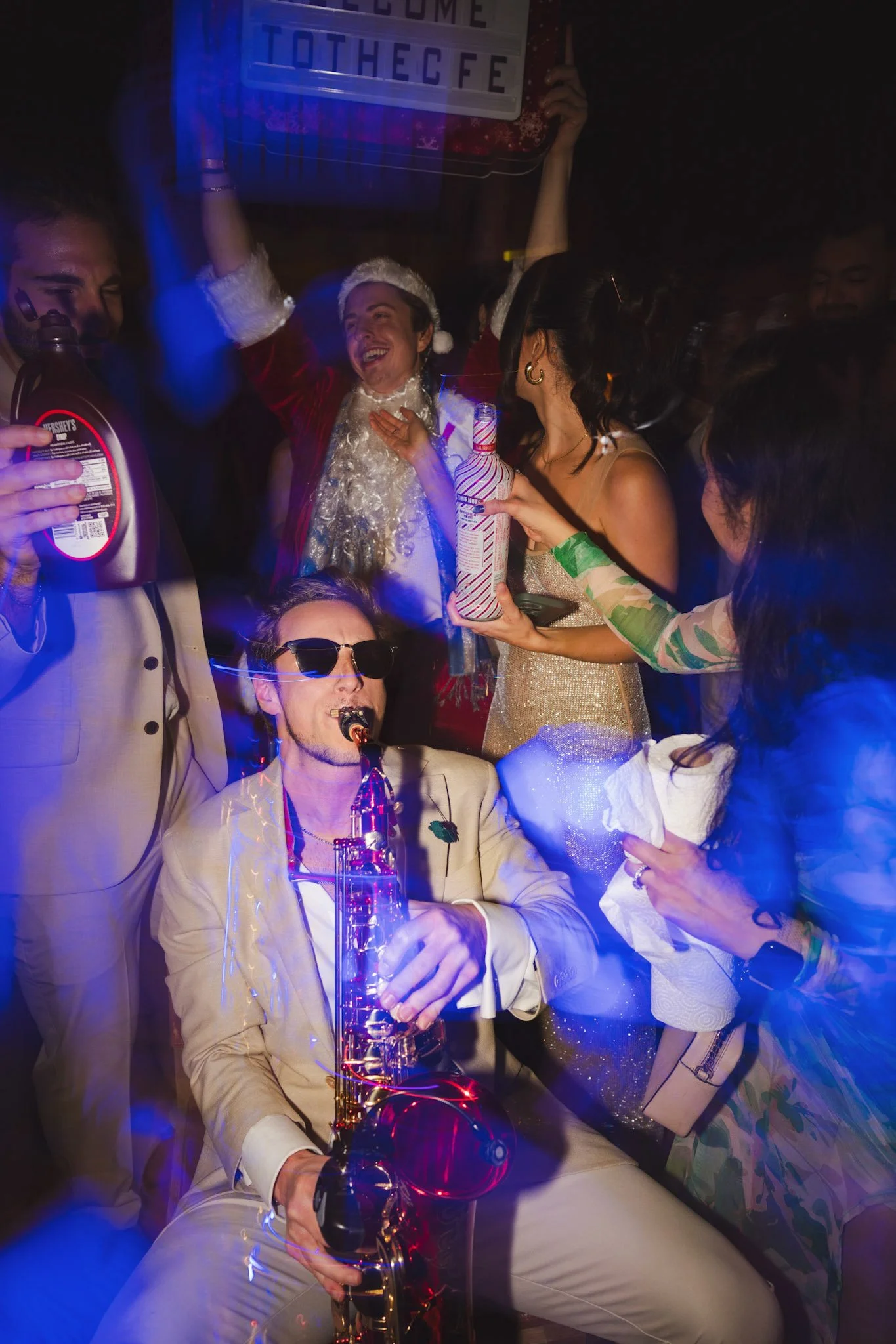 Christmas party people celebrating at a nightlife festival wearing festive attire, with Jason Nelson in a Santa hat and a woman with a microphone, in a dimly lit room with disco lighting.