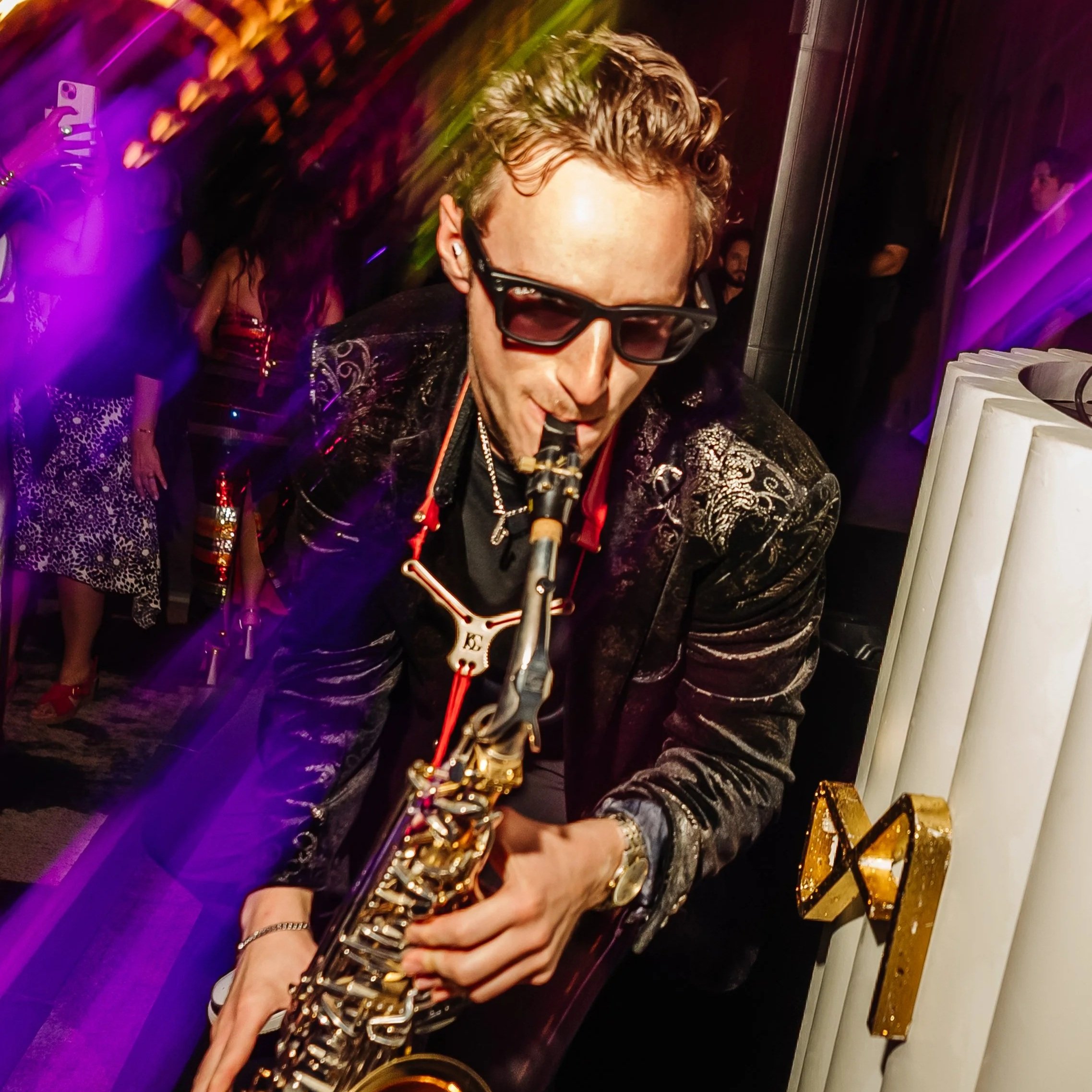 Jason Nelson wearing dark sunglasses and a black and gold blazer playing a saxophone at a corporate event with purple and gold decorations, and people dancing in the background.