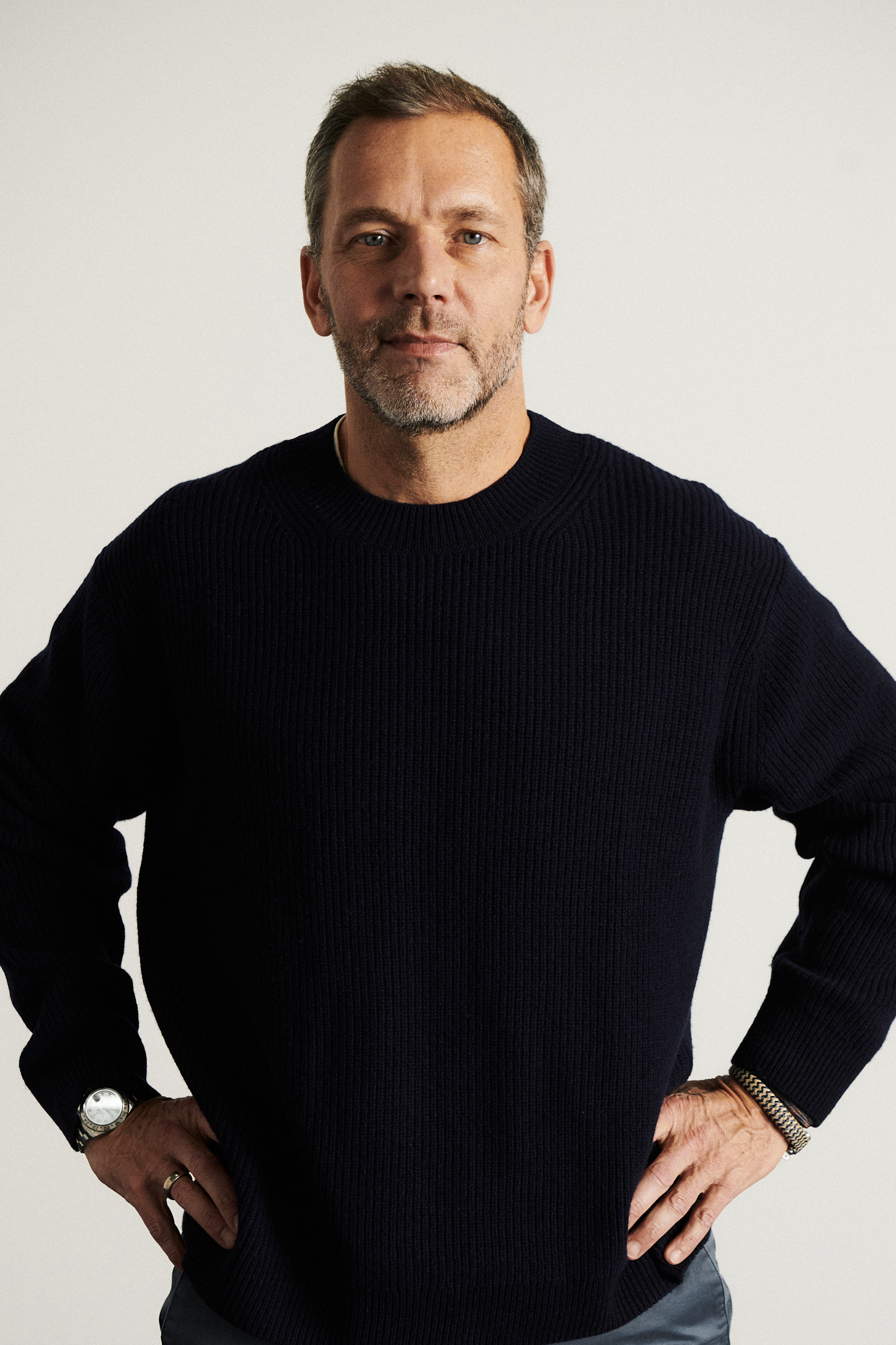 Middle-aged man with dark salt&pepper hair and beard wearing a navy blue sweater, with hands on hips, standing confident in front of a plain white background.