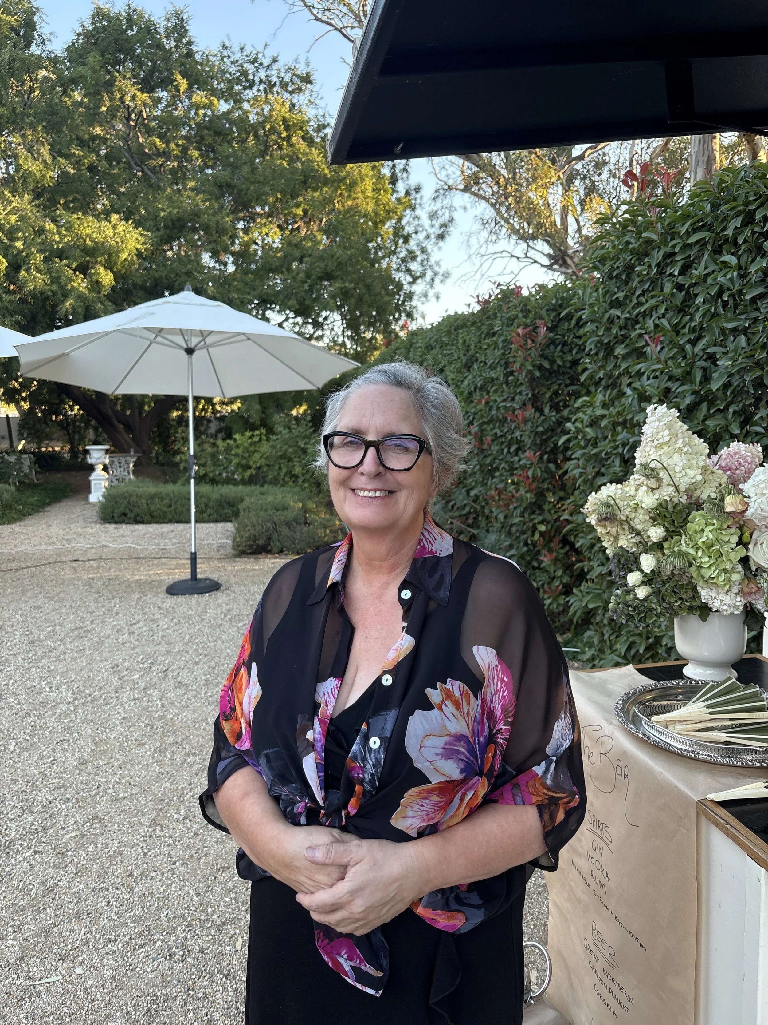 A woman smiling outdoors in front of greenery and a white umbrella, wearing a sheer black floral blouse.