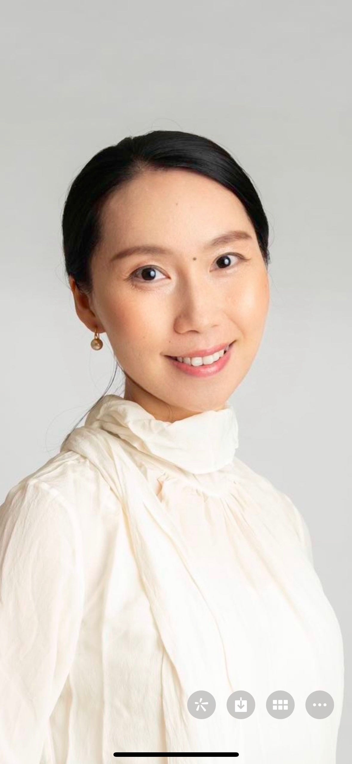 A portrait of a woman with black hair, wearing a cream-colored high-neck blouse and gold earrings, smiling against a plain light background.