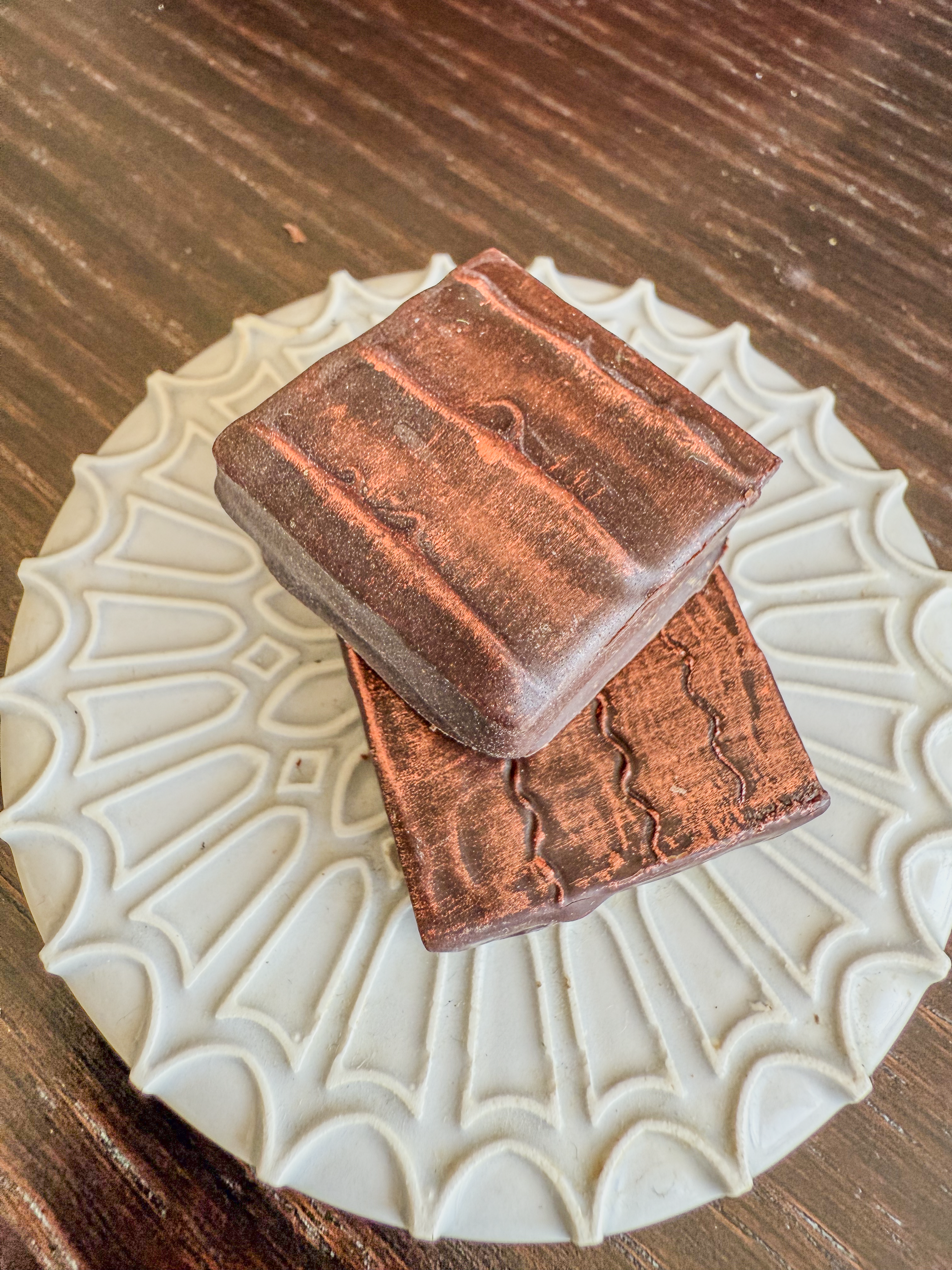 Two pieces of copper-colored soap with textured patterns on top, placed on a decorative white plate, on a wooden surface.