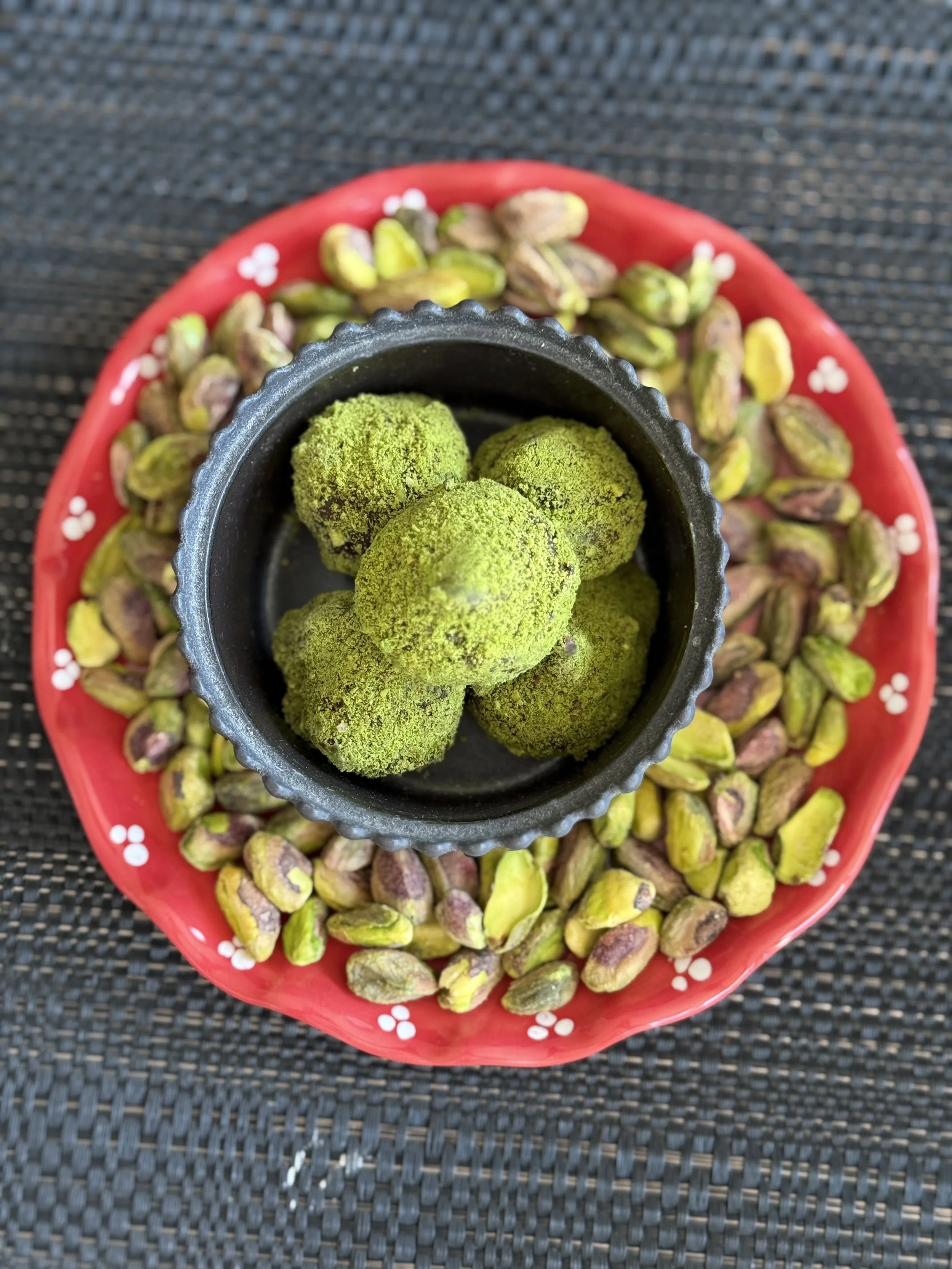 Green matcha-coated chocolate truffles in a black container, surrounded by roasted pistachios on a red dish with a flower pattern.