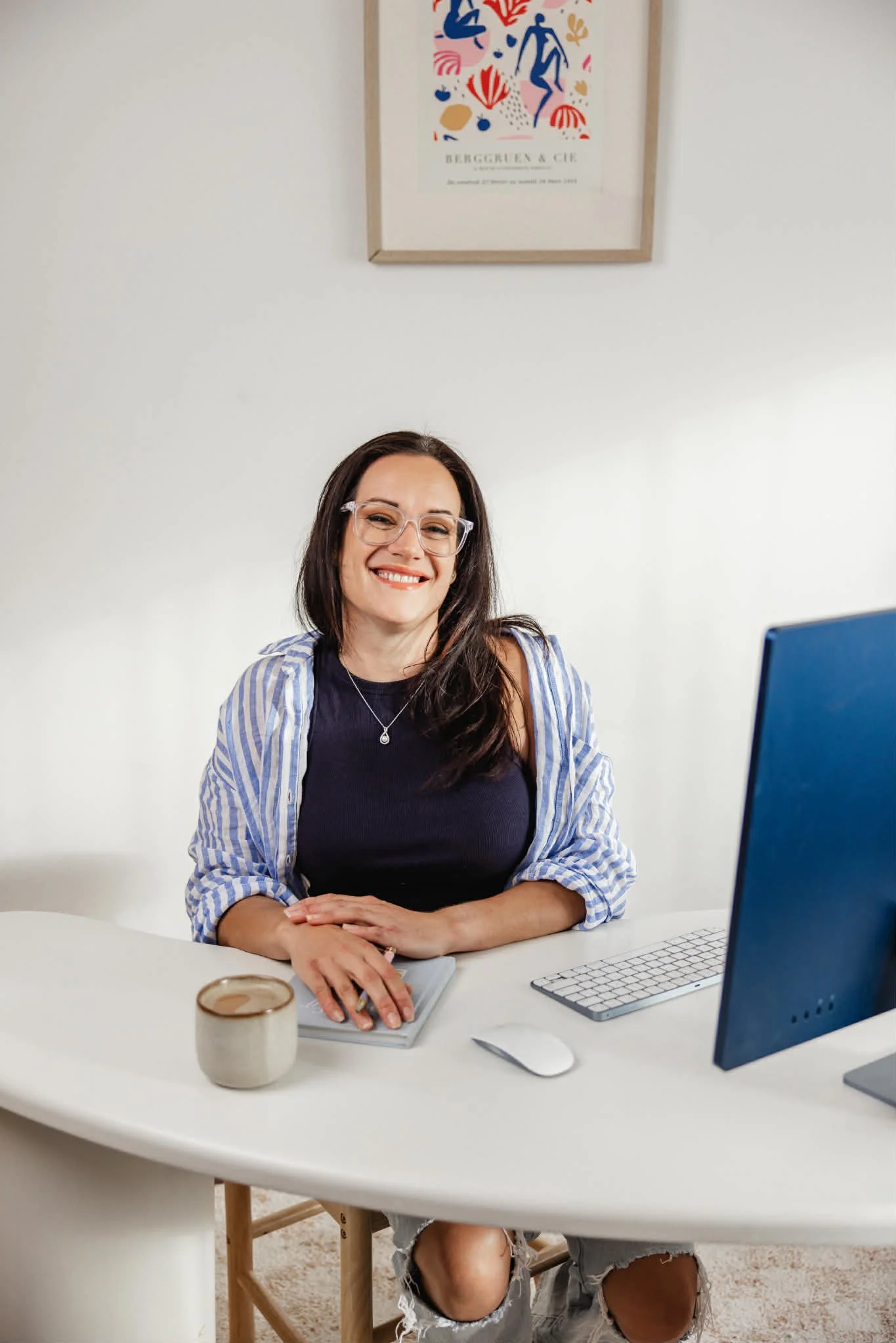 A woman sitting at a white desk with a computer monitor, keyboard, and mouse. She is smiling, wearing glasses, a black top, and a blue and white striped shirt. There is a coffee mug and a notebook on the desk. A framed abstract art piece hangs on the white wall behind her.