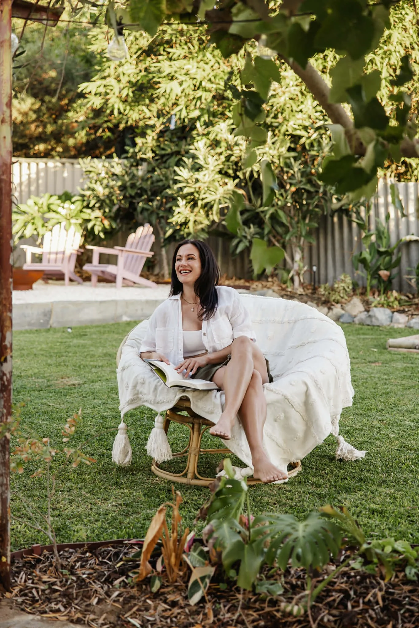 A woman sitting on a papasan chair with a blanket, reading a book in a garden with green grass and plants, and pink Adirondack chairs in the background.