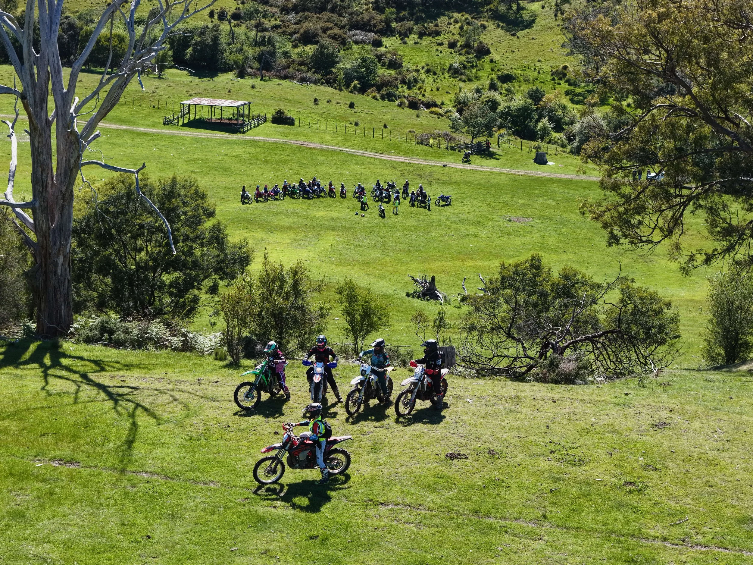 A group of motorcyclists gathered on a grassy hill, with some riding motorcycles and others standing. In the background, more motorcyclists are lined up on a flat field. The scene is set in a lush, green, hilly landscape with trees and a pathway.
