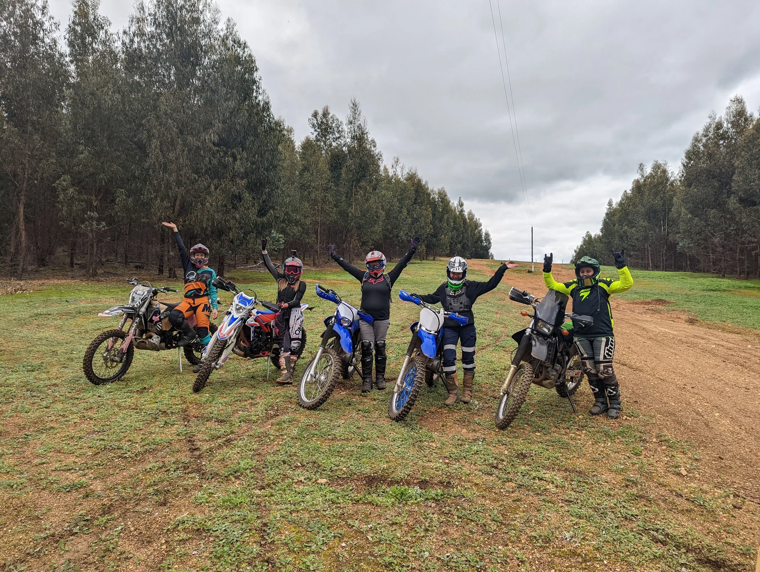 Five off-road motorcyclists standing next to their dirt bikes on a grassy trail with a forested area and cloudy sky in the background. They are wearing helmets, riding gear, and gloves, and are posing with arms raised.