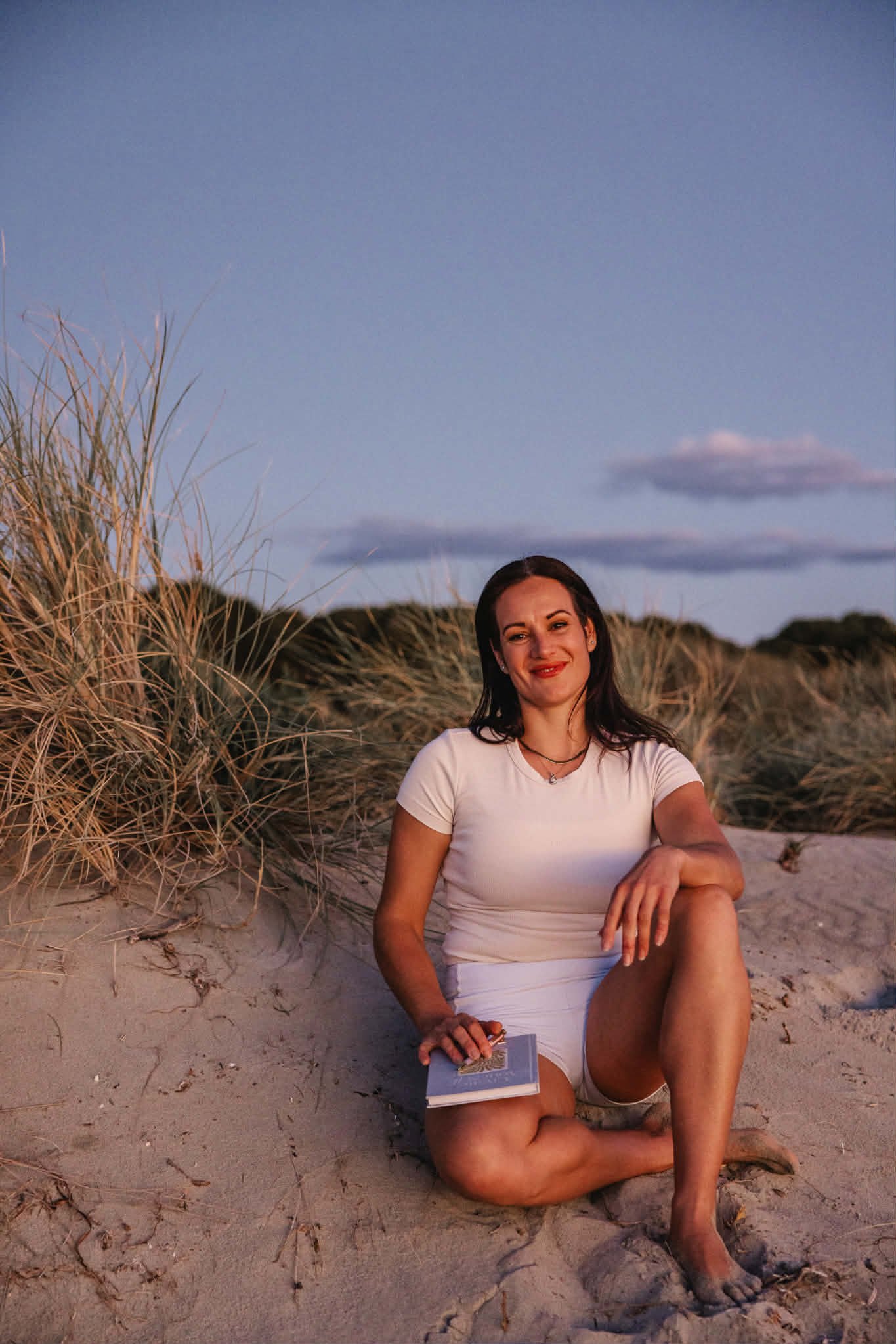A woman sitting on the sand at a beach during sunset, holding a book with one leg crossed over the other, with grassy dunes and a blue sky with a few clouds in the background.