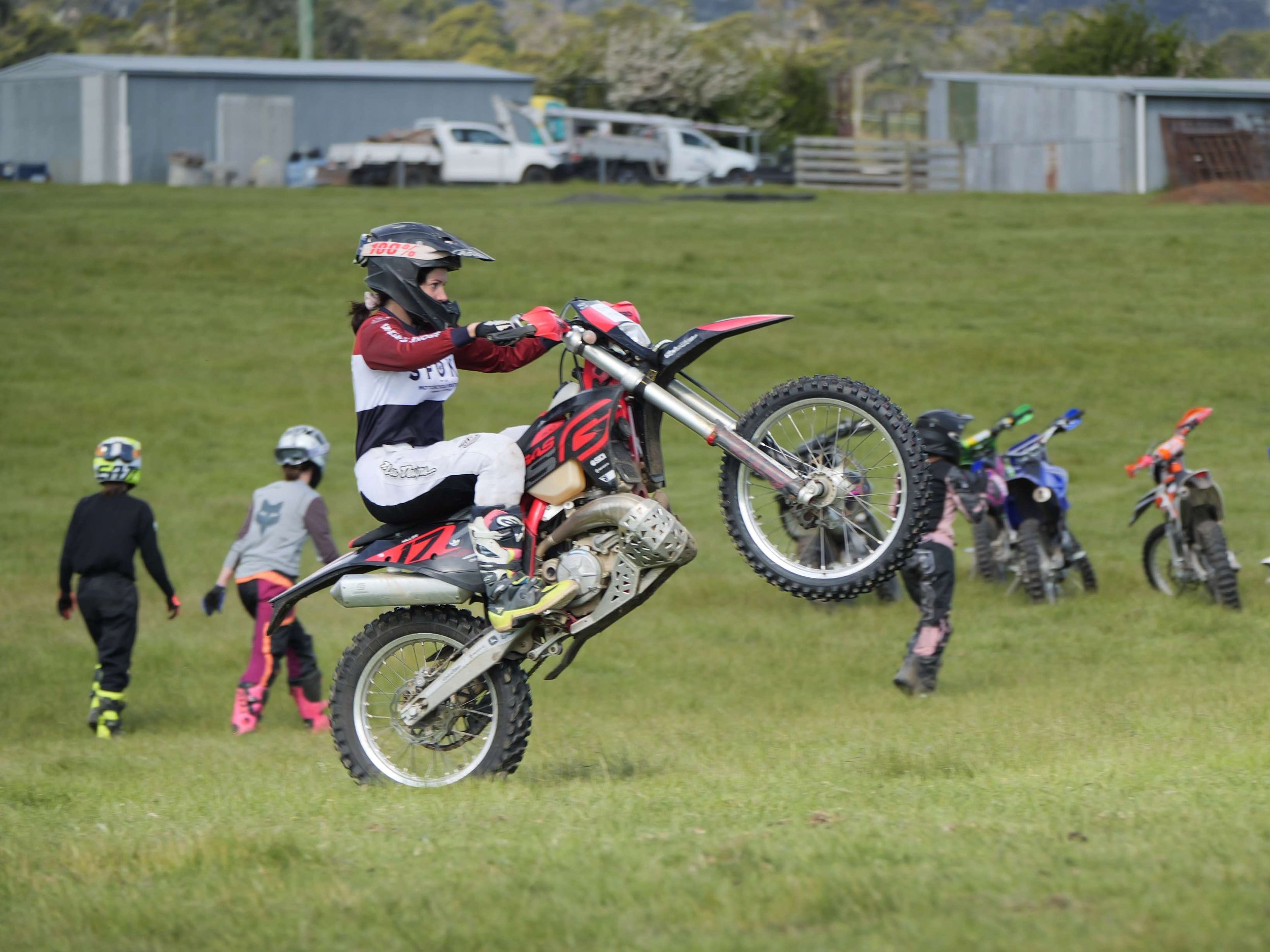 A female dirt bike coach in motocross gear performing wheelie on red and black dirt bike in green field, with other riders and sheds in background.