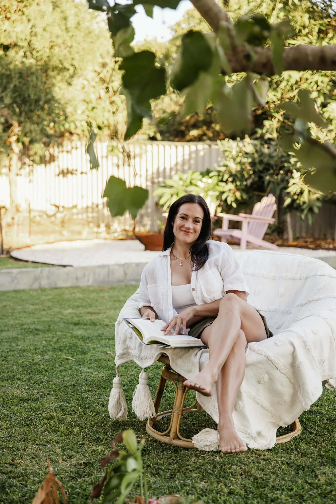 A female life coach sitting outdoors on a white cushioned chair, holding a book, smiling, with a green garden background and pink Adirondack chair in the background.