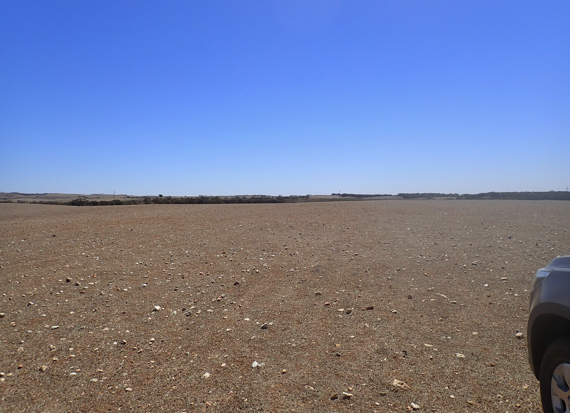 Revera Energy Robertstown Project image of a vast, empty desert landscape with sandy, rocky terrain and a clear, bright blue sky. A partial view of a gray car is visible on the right side of the image.