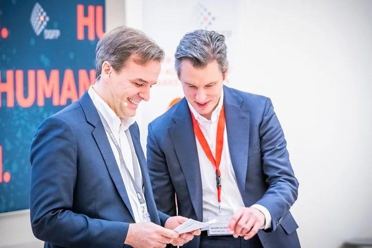 Jochen Menges standing with another man in suits, smiling and looking at a document together during a conference.