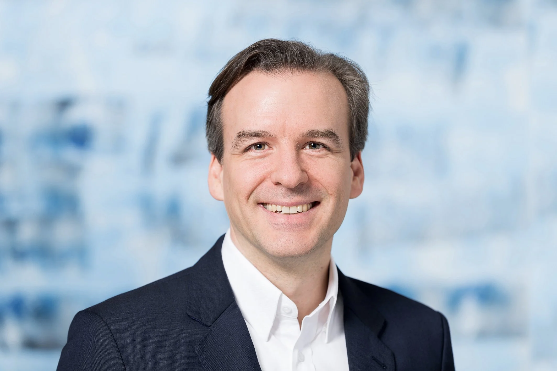Portrait of Jochen Menges, smiling in a dark suit jacket and white shirt against a blue blurred background.