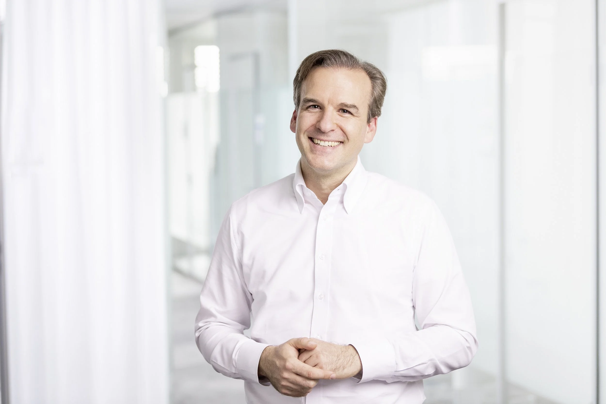 Jochen Menges smiling with his hands clasped in an a bright office background.