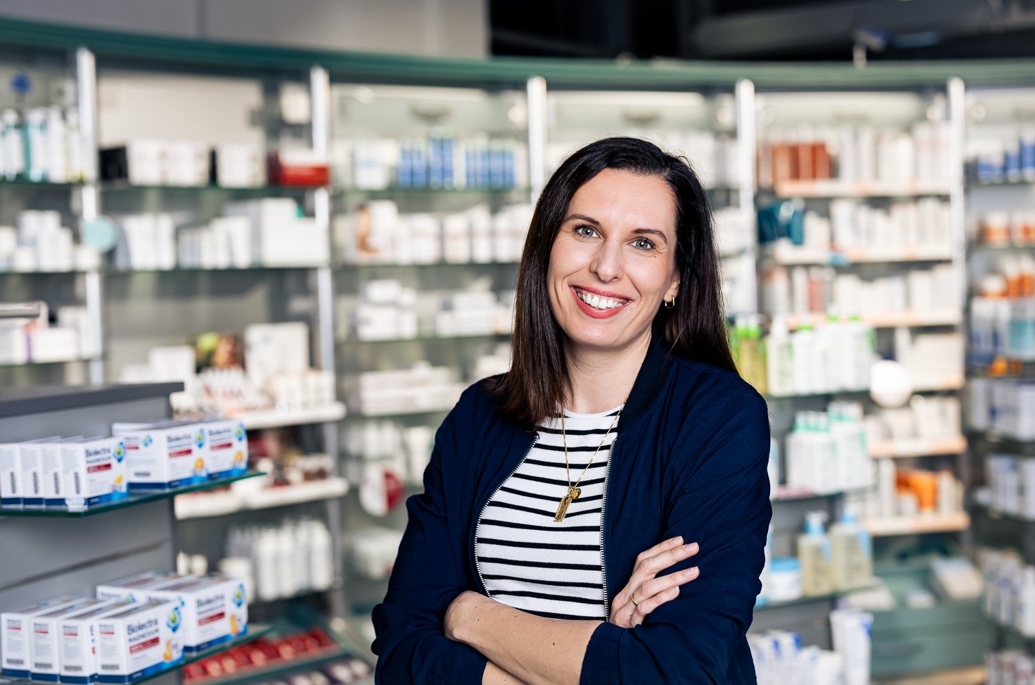 Eine lächelnde Frau mit dunklen Haaren, trägt eine schwarze Jacke und gestreiftes Top, steht in einer Apotheke vor Regalen mit Medikamenten.