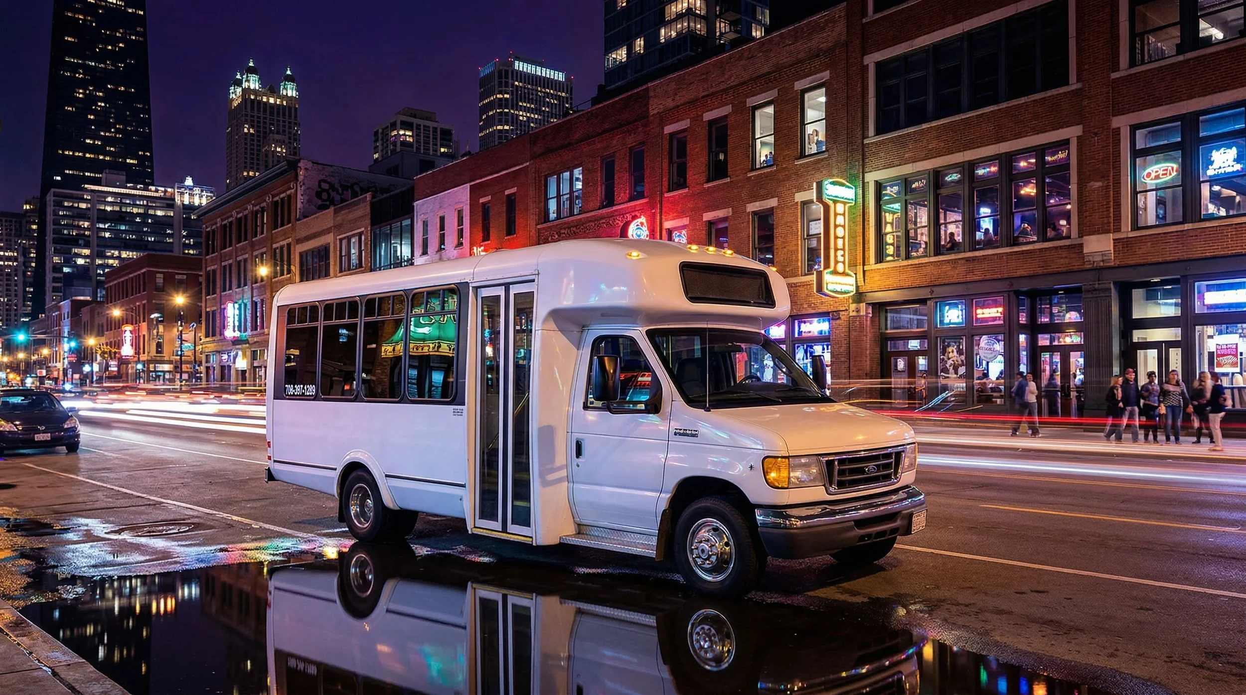 Nighttime city street scene with a white shuttle bus parked in the foreground, reflecting in a large puddle. Behind, a brick building with neon signs and windows occupied by people. Tall skyscrapers with lit windows are visible in the background.