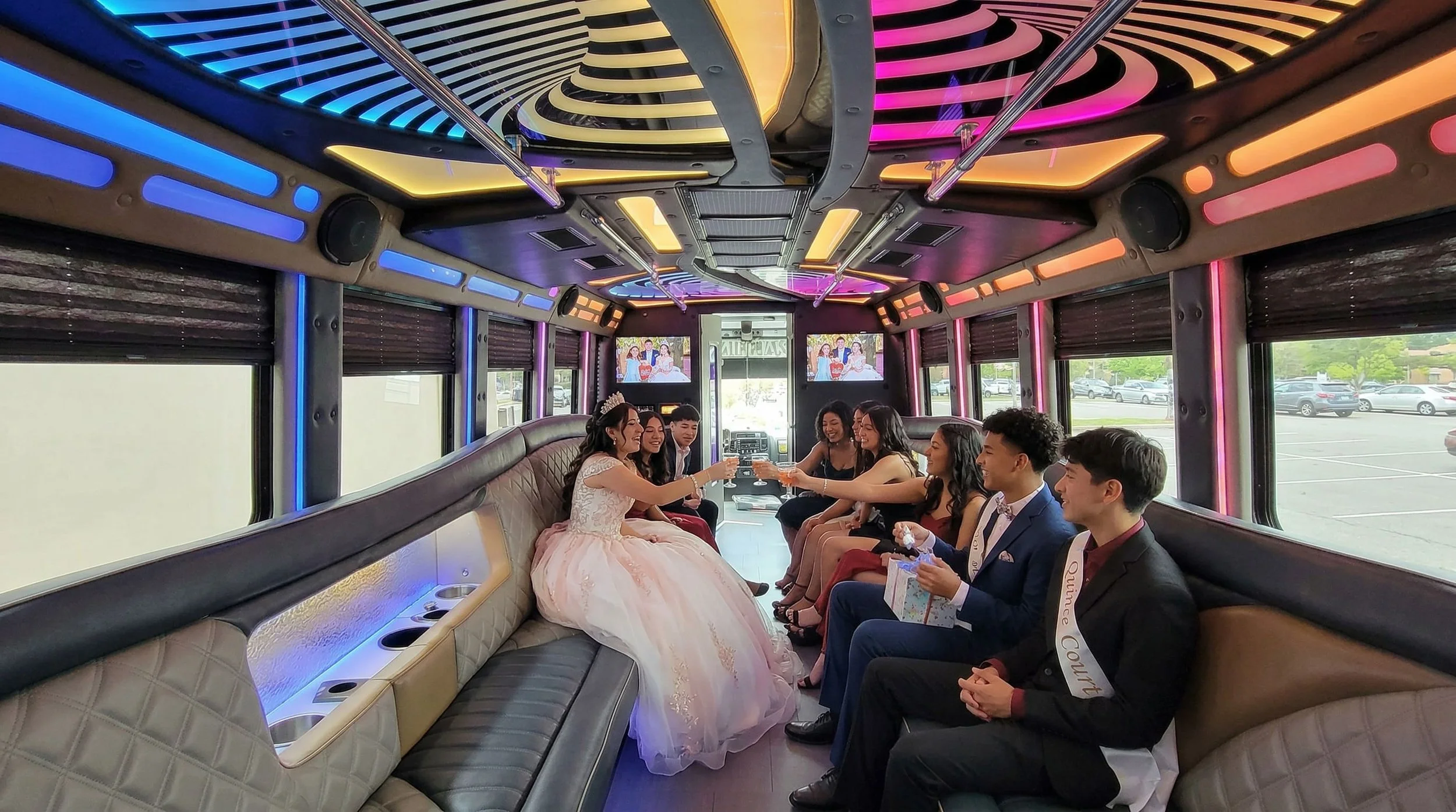 People celebrating a Quinceañera inside a decorated party bus with colorful LED lights and large windows, some holding glasses, with a woman in a pink gown and tiara.