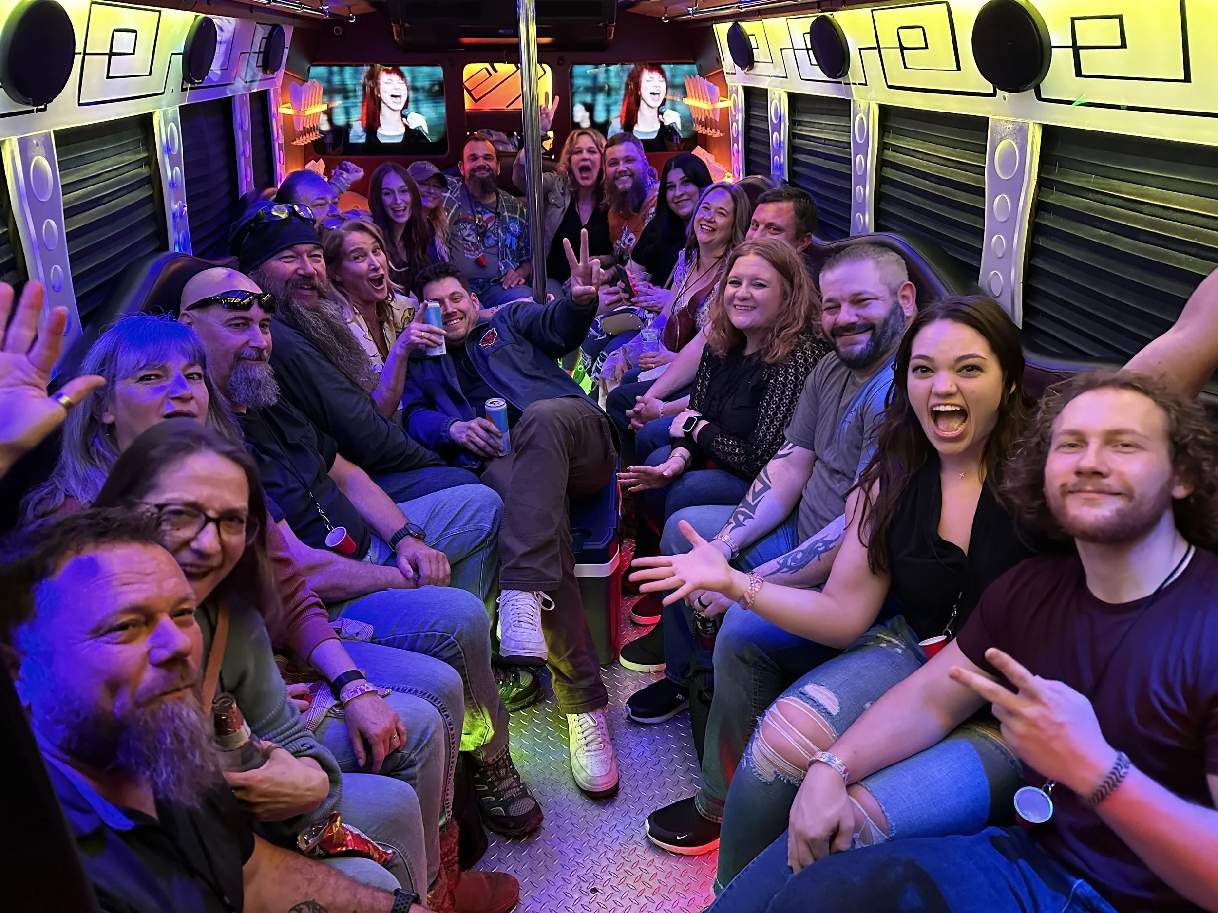 A group of people inside a party bus, smiling and celebrating, with colorful lighting and a large screen at the front showing a woman singing.