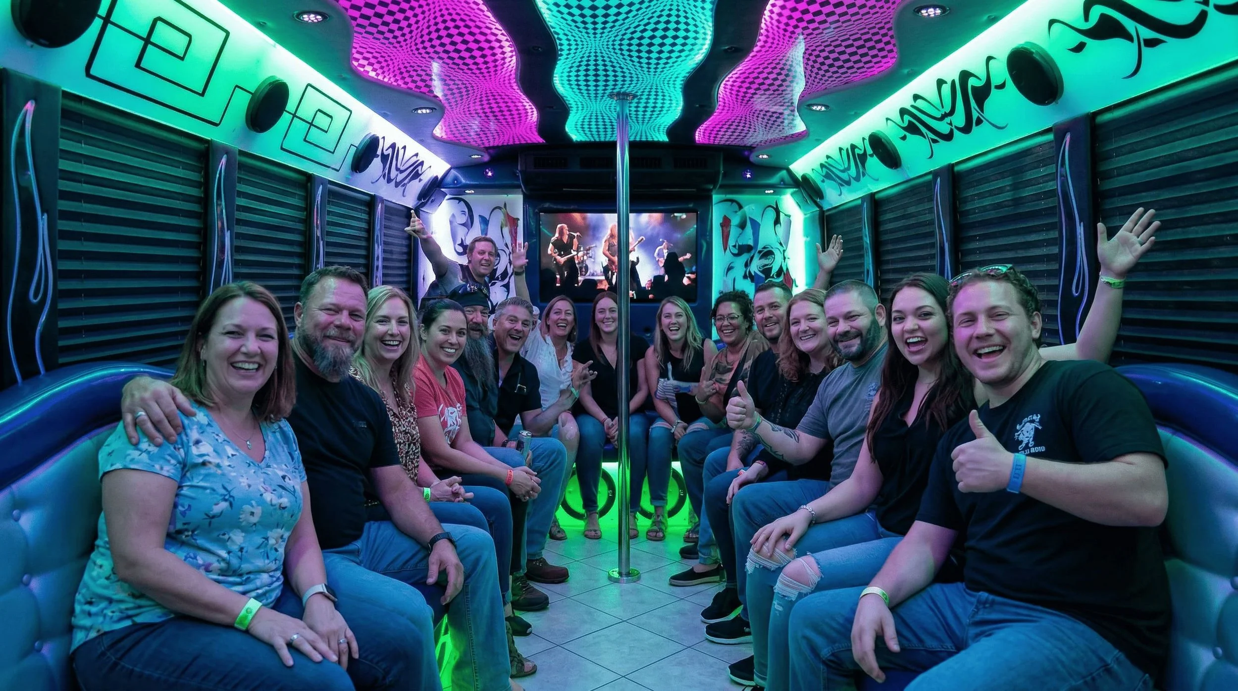 A group of people sitting inside a colorful party bus with vibrant neon lighting, a pole in the center, and a digital screen showing a live band performance.