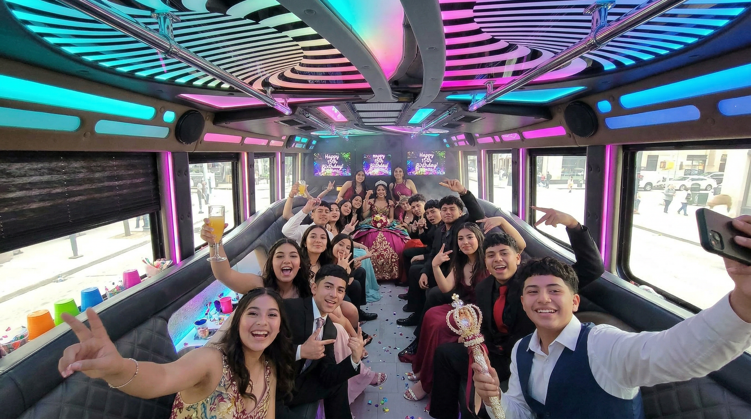 A group of young people celebrating a 15th birthday inside a decorated party bus. The birthday girl is wearing a traditional dress and tiara, surrounded by friends making peace signs and smiling. There are colorful LED lights on the bus ceiling and screens in the background that read 'Happy 15th Birthday'.