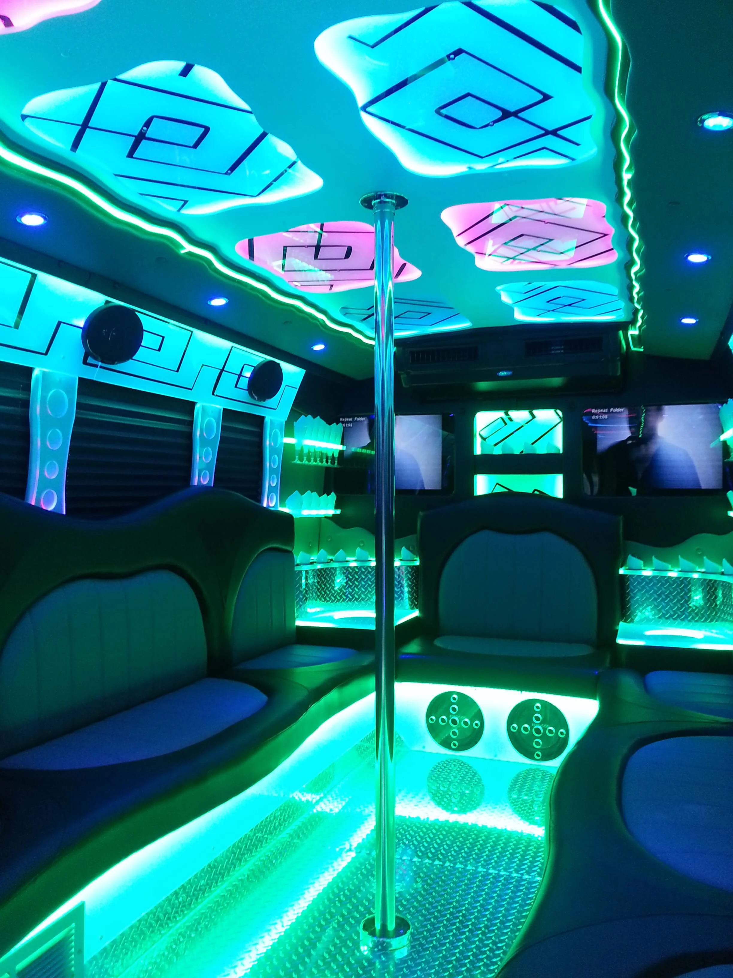 Interior of a dimly lit karaoke room with neon green, blue, and pink lighting, featuring a pole in the center, curved booth seating, and decorative illuminated panels with geometric patterns.