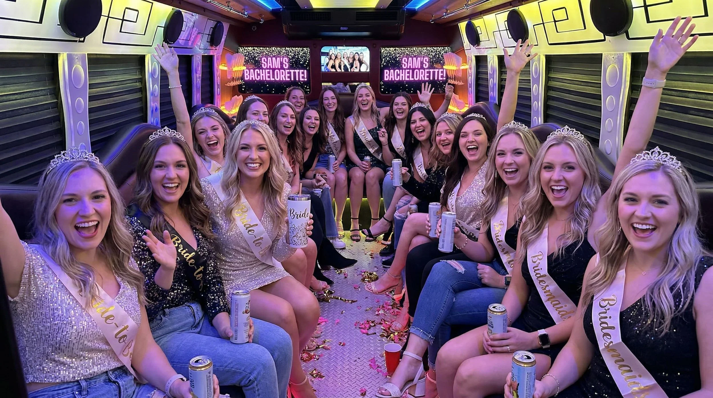 Group of women wearing pageant sashes and tiaras celebrating during a bachelorette party inside a decorated party bus, with a screen that reads 'Sam's Bachelorette!'