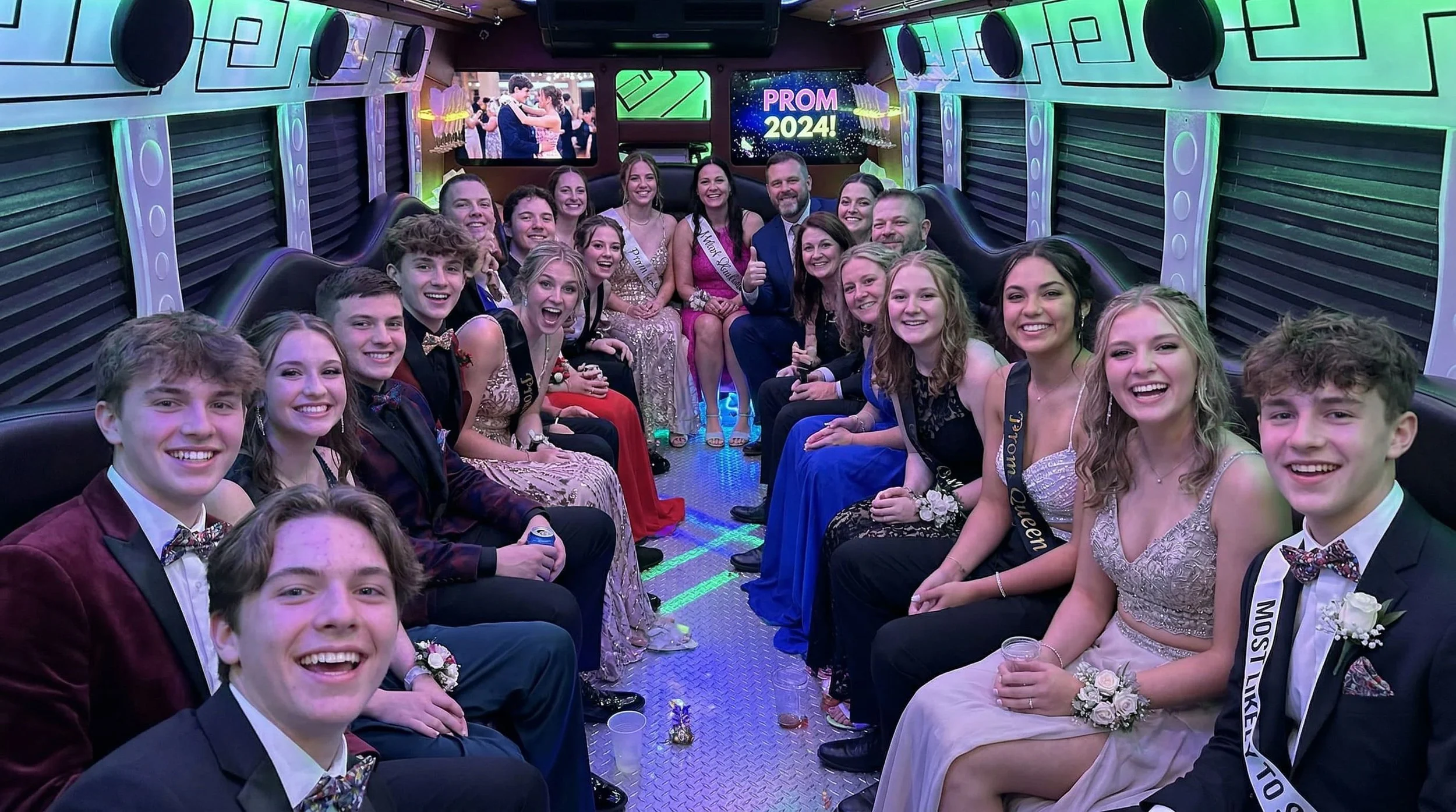 A large group of teenagers dressed in formal attire inside a decorated party bus with neon lights. The teenagers are smiling and posing for the camera, and the background on a screen reads "PROM 2024!"
