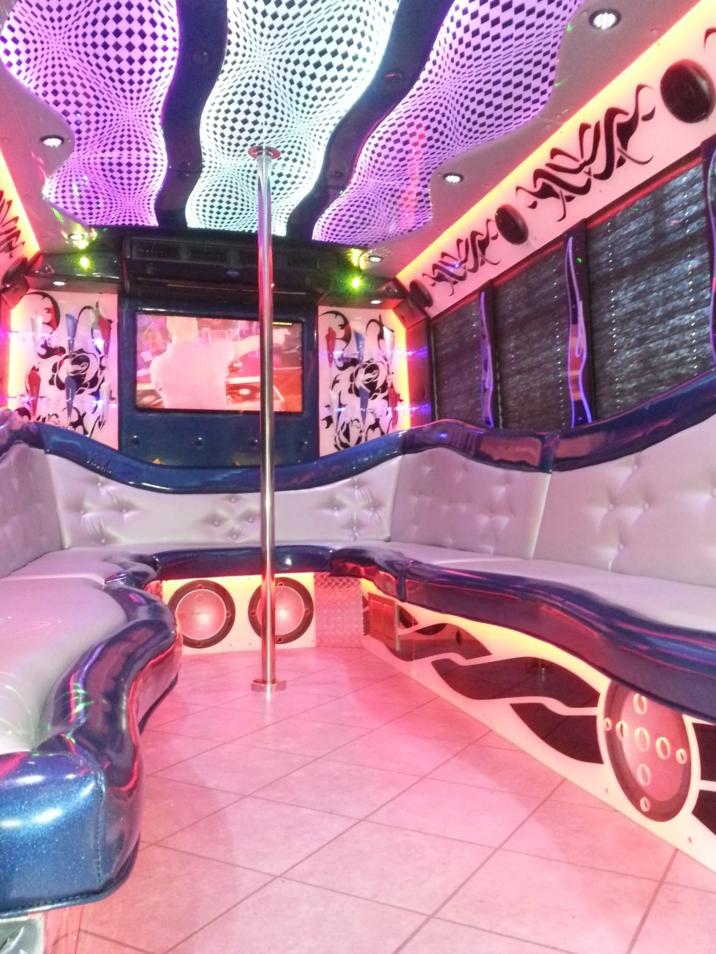 Indoor entertainment room with a pole in the center, illuminated ceiling with black and white swirling patterns, pink and purple lighting, padded seating, and a musical clock on the wall.