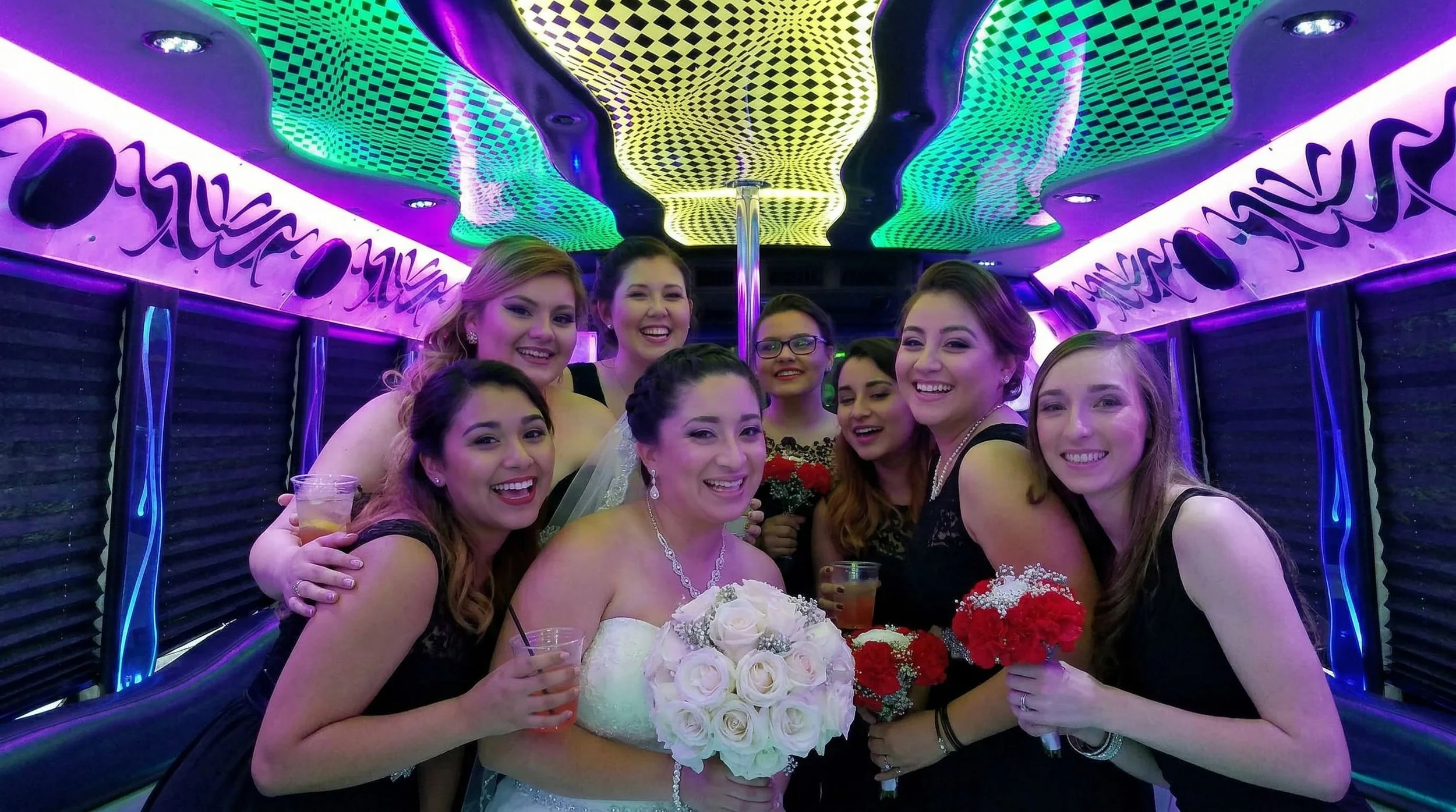 Group of women celebrating a wedding, with a bride holding a bouquet of white roses, in a room with colorful, neon lighting.