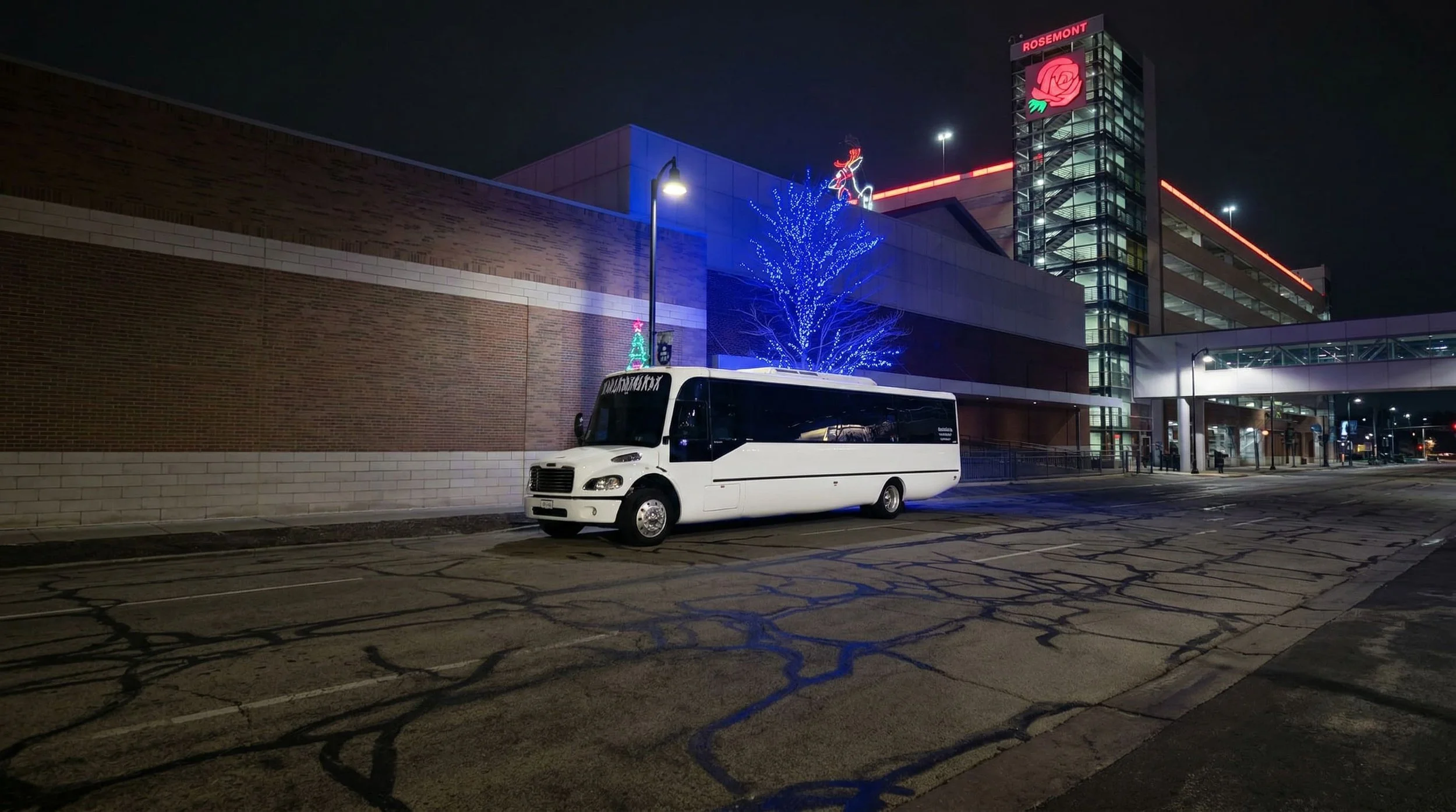 Empty parking lot in front of a modern building at night, decorated with blue Christmas lights on a tree, illuminated reindeer and Christmas figures on the building, and a white bus parked near the sidewalk.