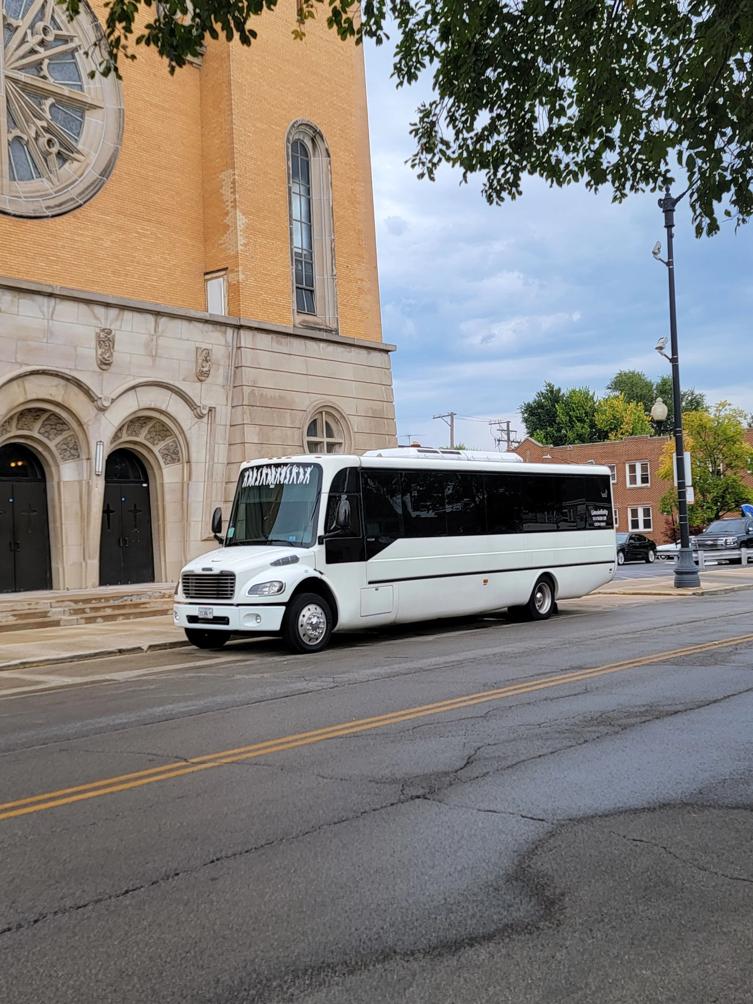 A white and black shuttle bus parked on the street next to a brick church with arched windows and a large clock on the tower. The street has yellow double lines, and there are trees and other buildings in the background.