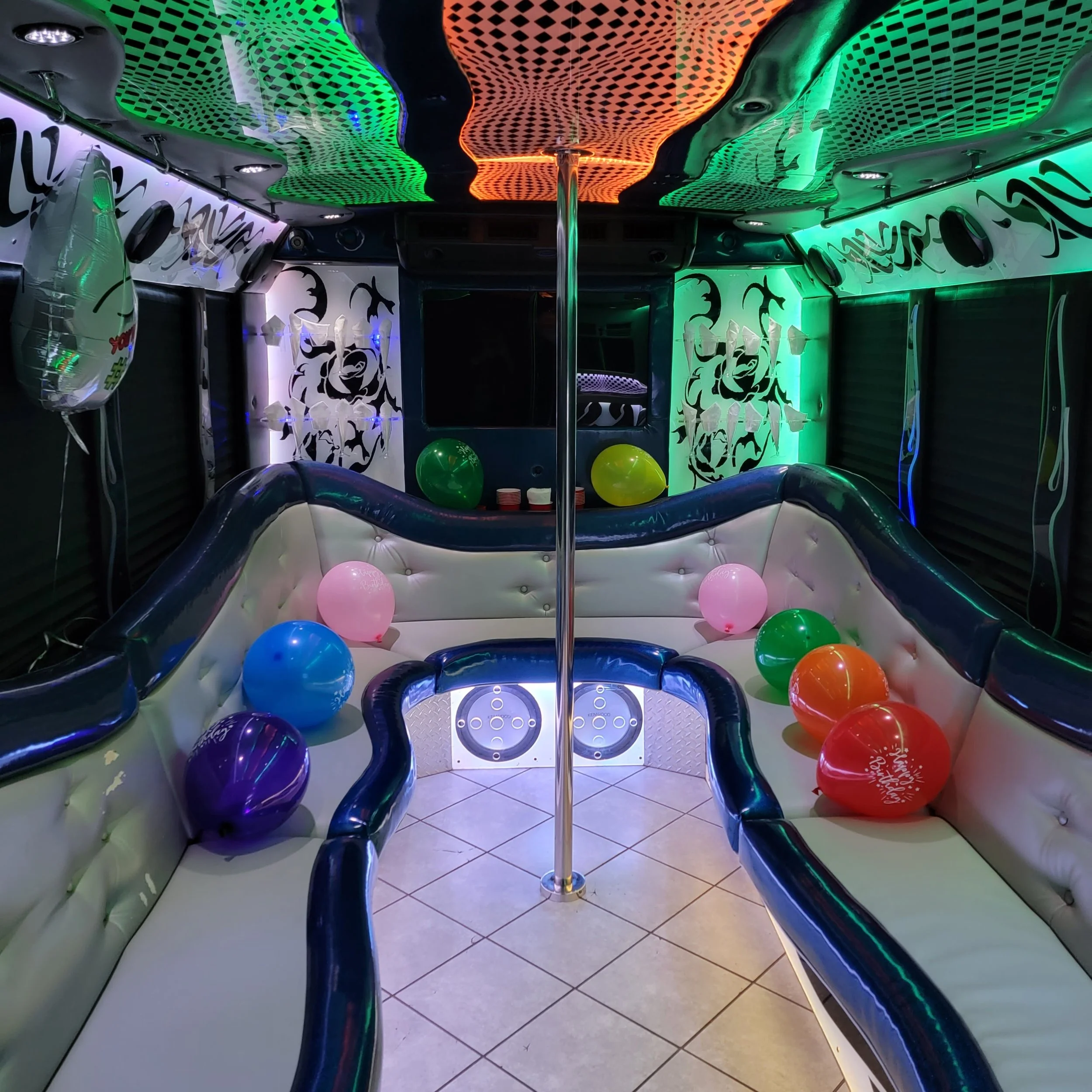 Interior of a party bus with black, green, orange, and blue LED lighting, decorated with balloons, a pole in the center, and black and white patterned walls.